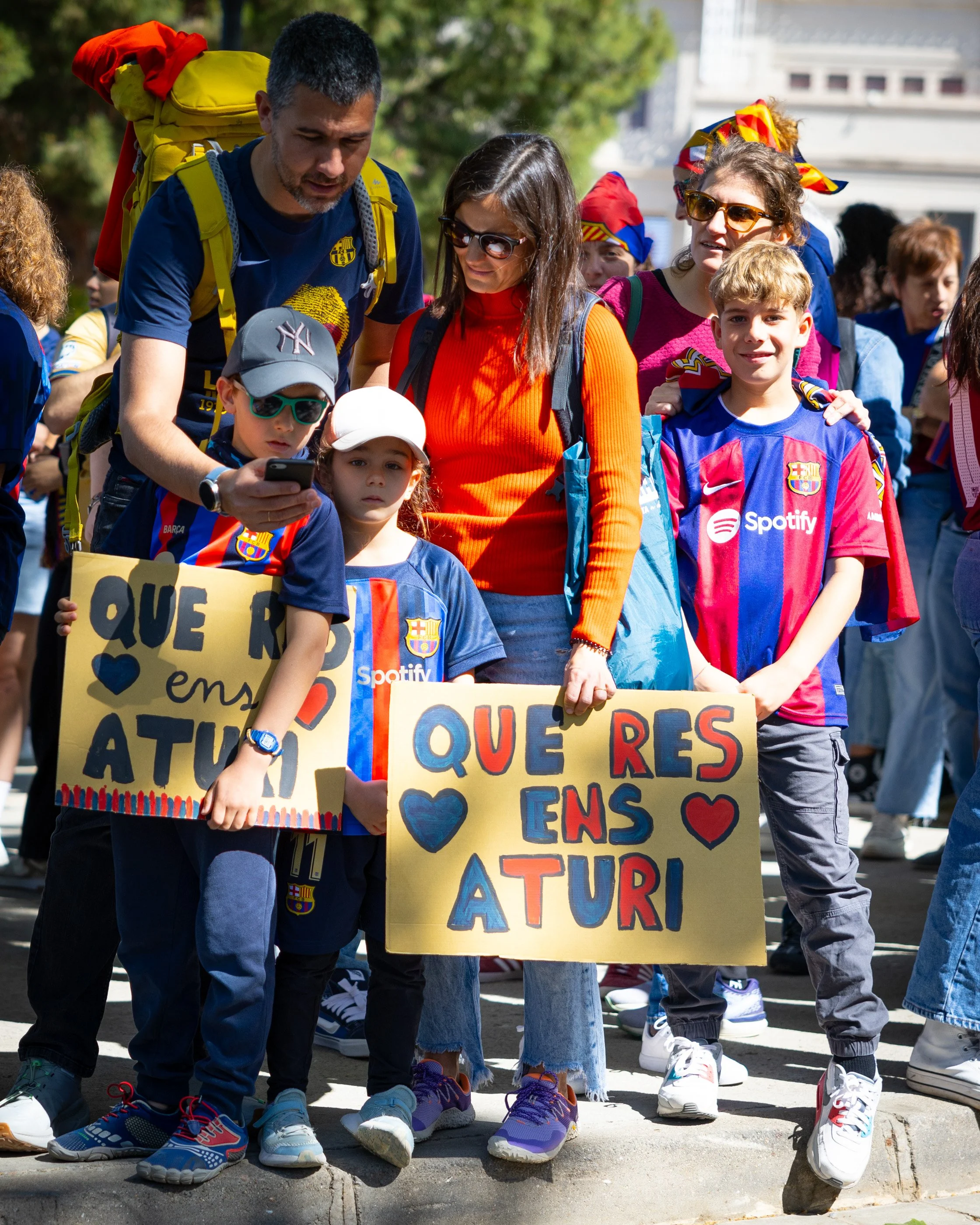 A group of children and a man gathering together outdoors, holding signs with messages in Spanish, in a city setting with trees and buildings in the background.