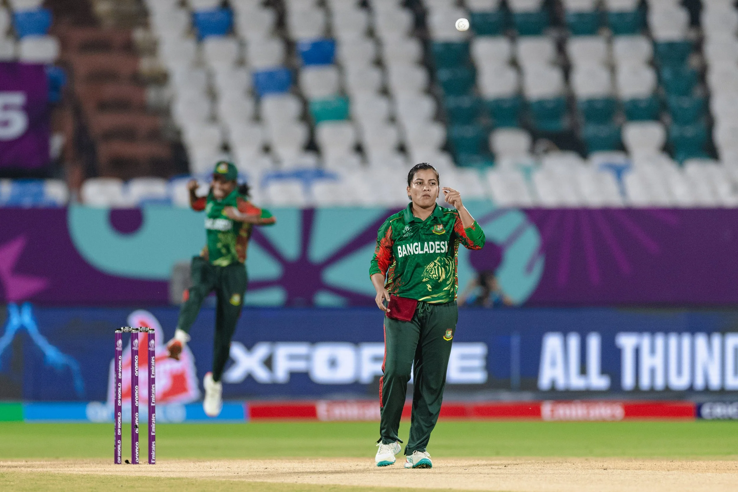 A female cricket player from Bangladesh on the field during a match, wearing a green and red team uniform, with a woman in the background also in uniform. The field has stumps and pitch markings, with empty stadium seats and advertisements visible.