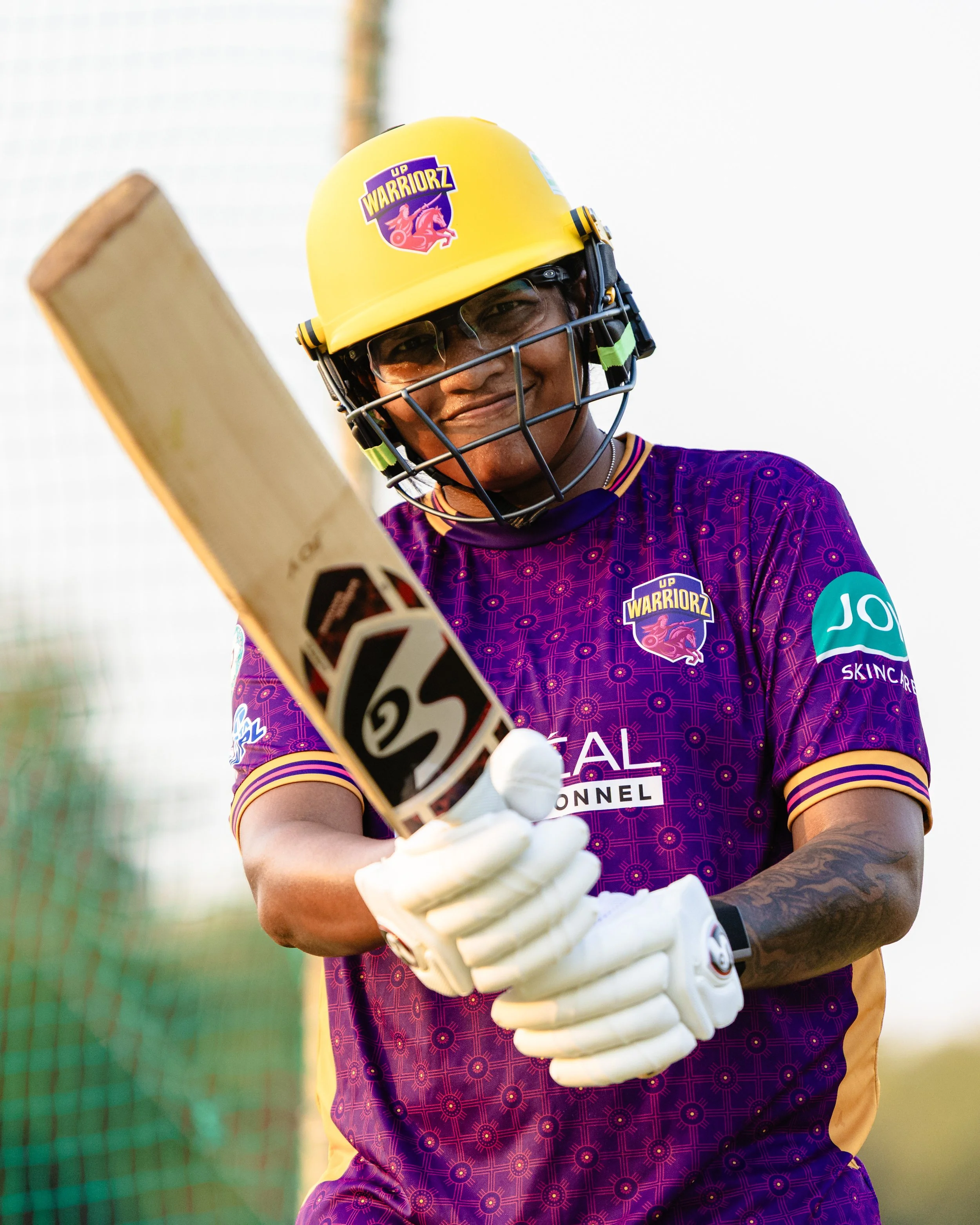 A person in a purple sports jersey and yellow helmet with a logo that says 'UP WARRIORZ' hold a cricket bat. The person is wearing glasses and white gloves. The background is blurred.
