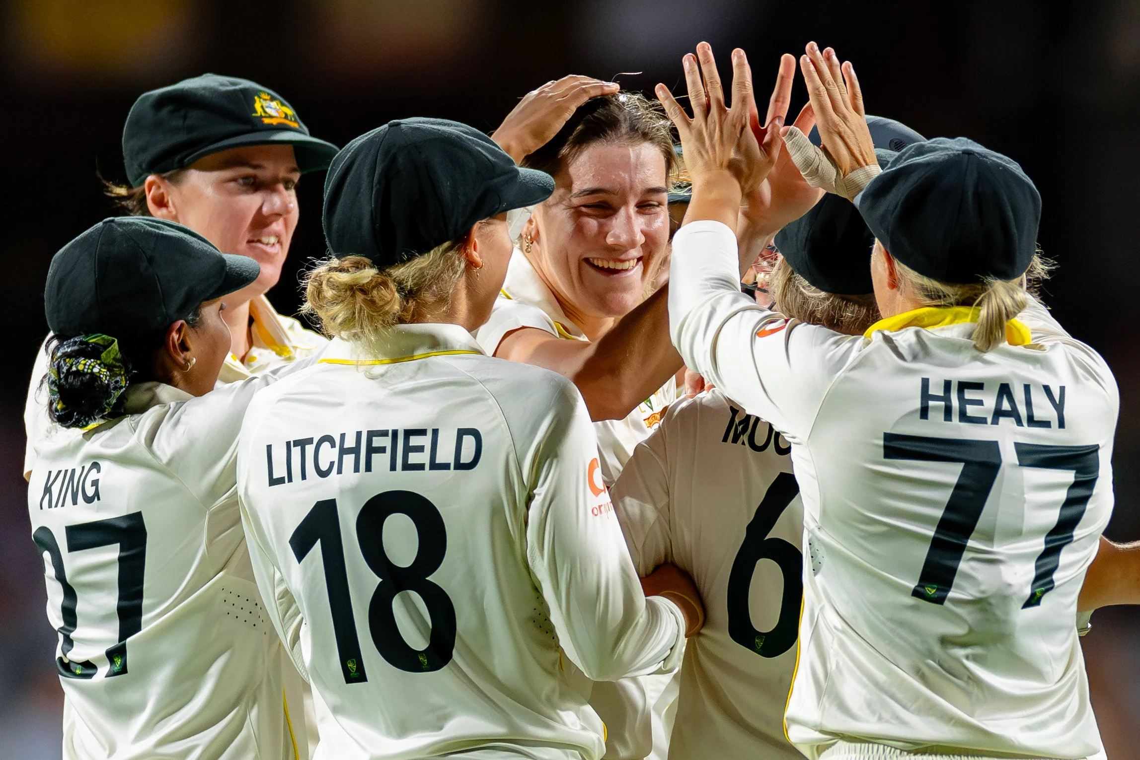 A team of female cricket players celebrating on the field, with some applauding and smiling, wearing white jerseys with black numbers and names, black caps, and some with yellow scarves.