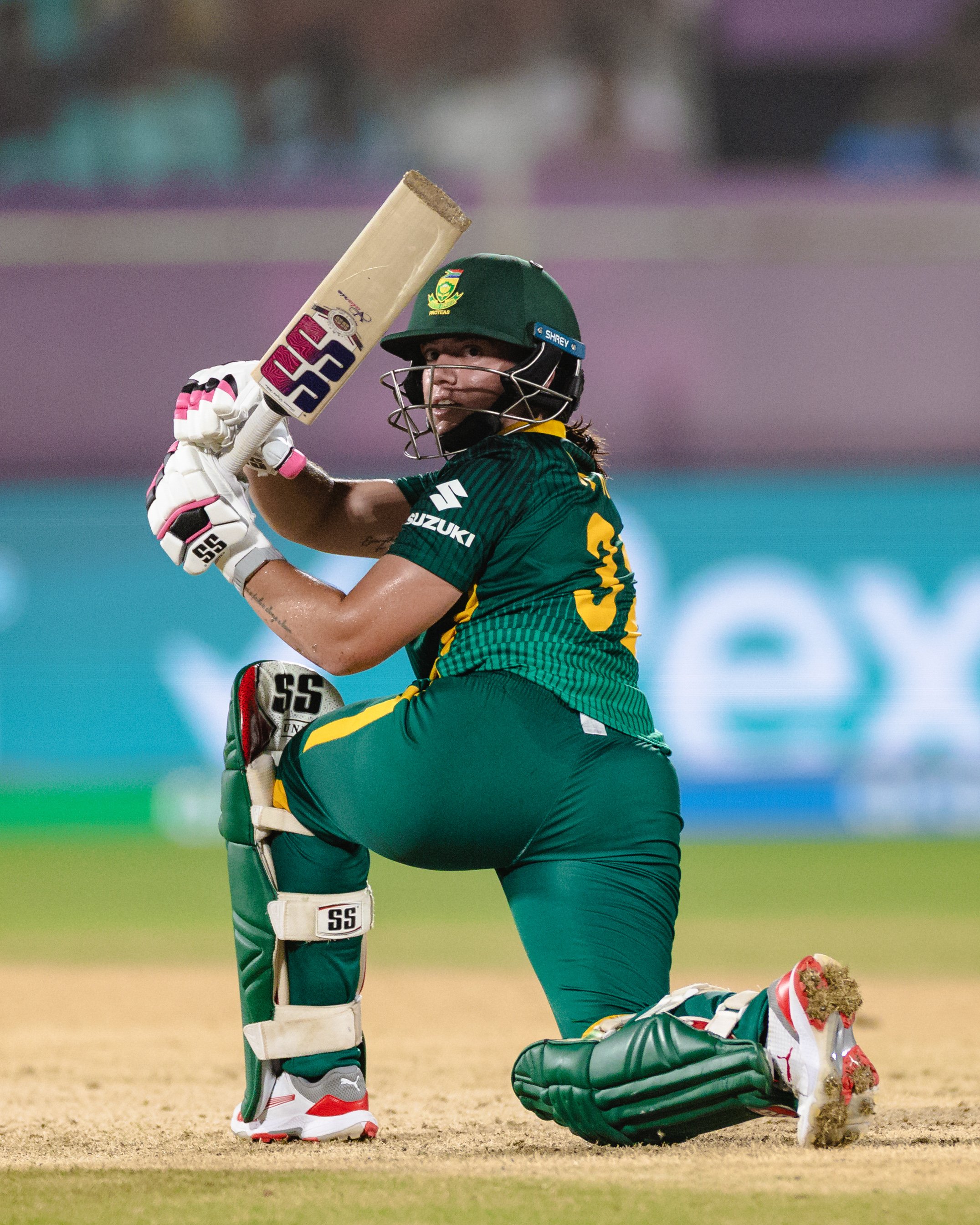A cricket player wearing green and yellow uniform, kneeling on the ground with a cricket bat, helmet, gloves, and pads, looking at the camera.