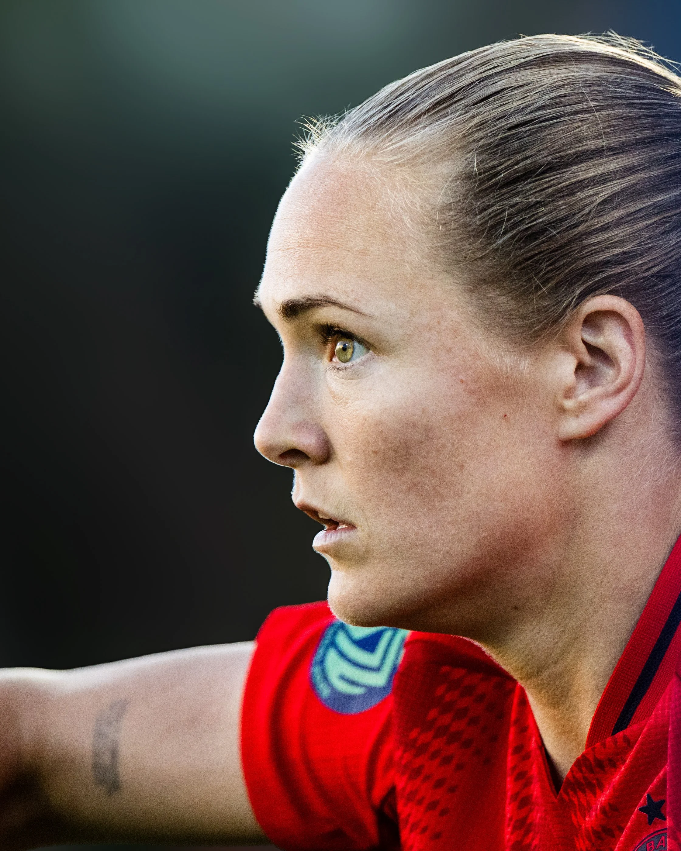 Close-up of a young female athlete with blonde hair, wearing a red sports jersey with blue and black details, focused and looking to the side, with a blurred background.