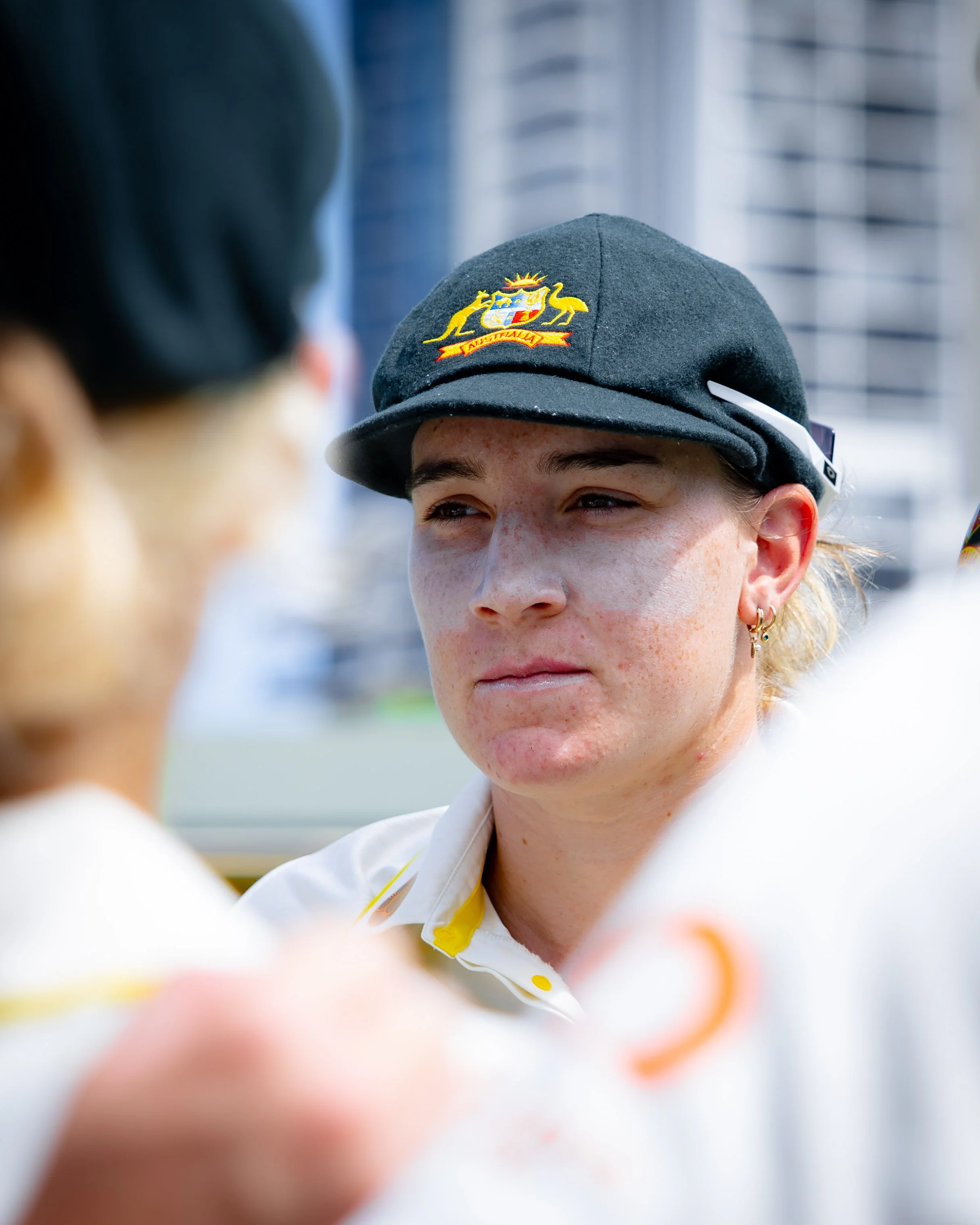 Close-up of a young person wearing a gray hat with an embossed emblem, smiling with slightly squinted eyes, and wearing a white shirt with yellow accents, outdoors in a city environment.
