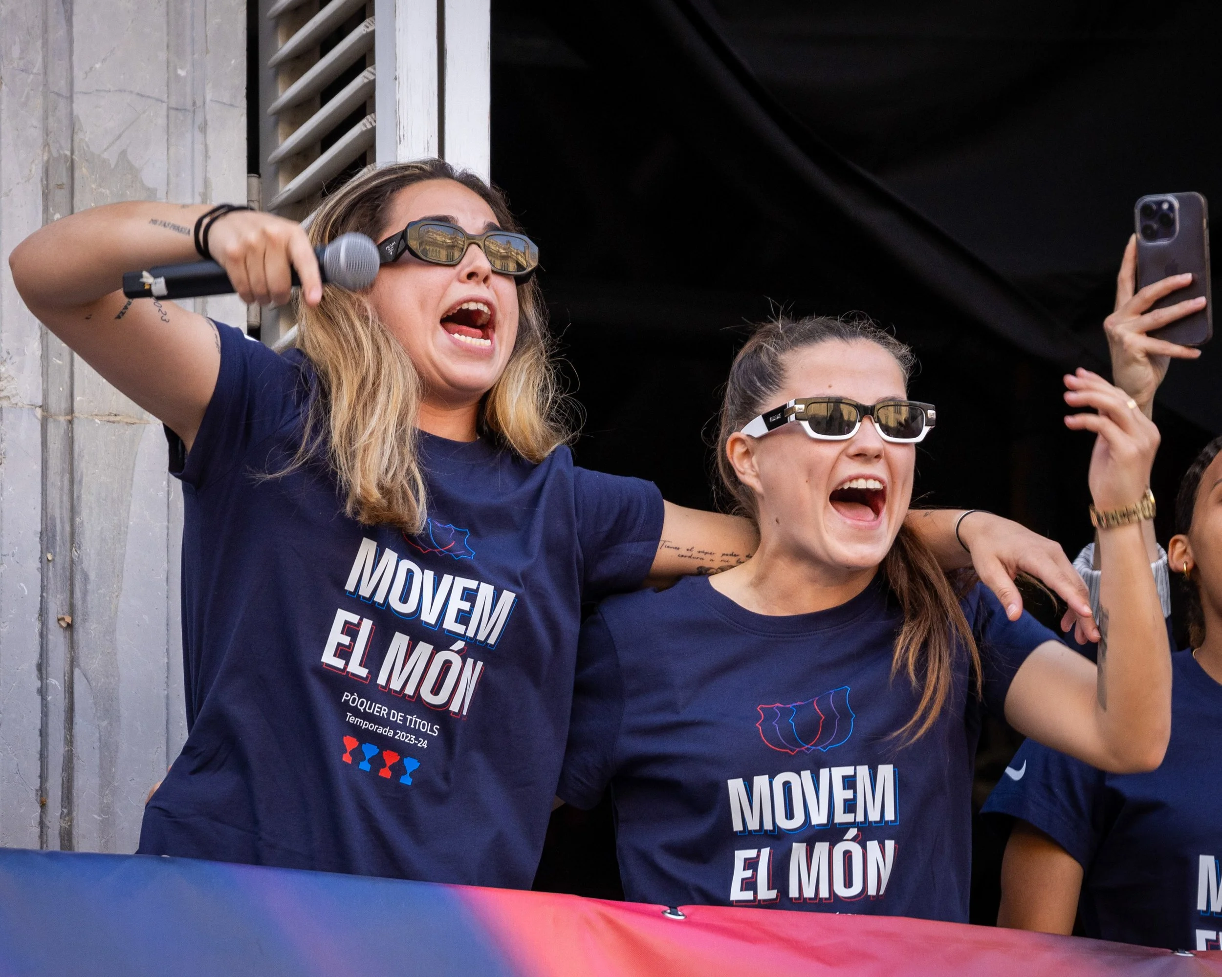 Group of women wearing dark blue shirts with political message, standing together with arms around each other, singing or shouting, holding a microphone and a smartphone, with a banner in front of them.