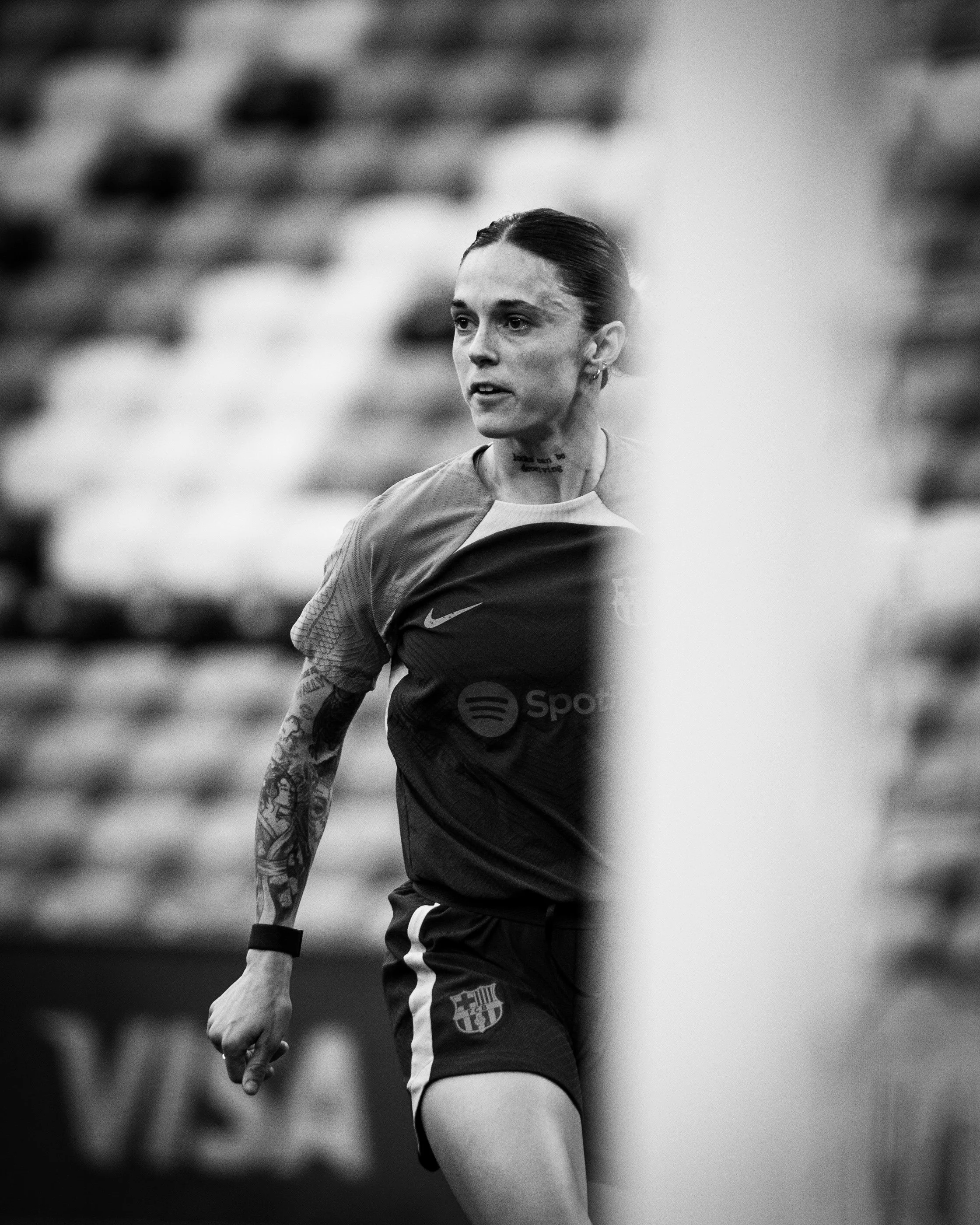 A female athlete with dark hair tied back, tattoos on her arm, wearing a sports jersey with the FC Barcelona logo and Nike logo, standing outdoors with a blurred background of bleachers or seats.