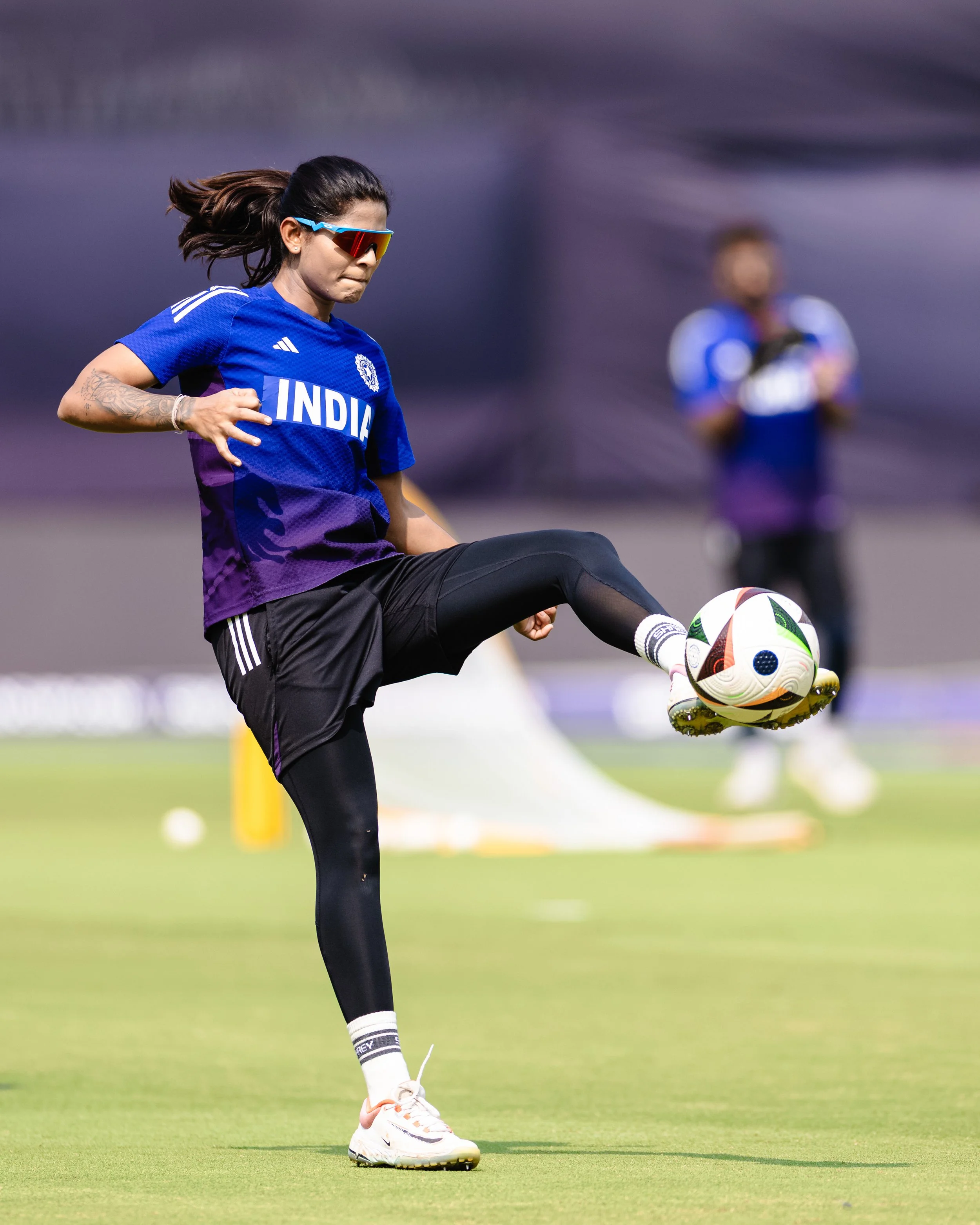 A female soccer player wearing sunglasses and a blue India jersey practicing a kick with a soccer ball on the field.