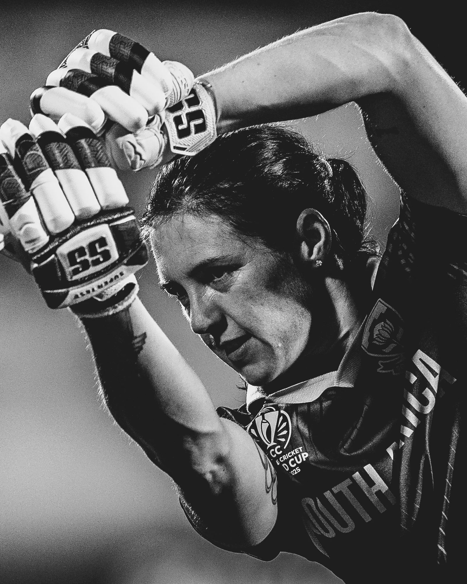 A female athlete in a cricket uniform, wearing gloves, with her hair tied back, highlighting her face and part of her uniform, in a black and white photo.
