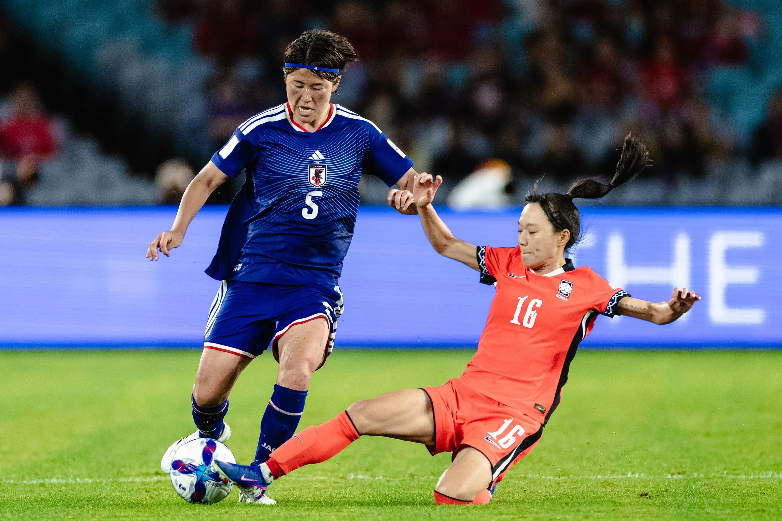 Two female soccer players, one in a blue uniform and one in a red uniform, competing for the ball on a green field, with a blurred crowd in the background.
