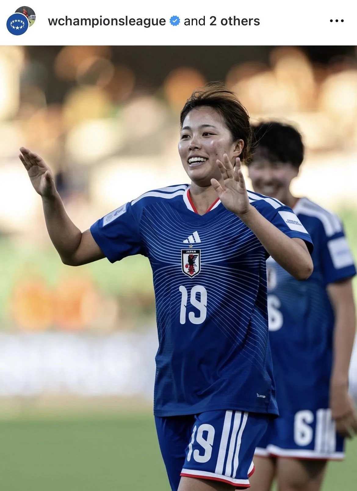Japanese female soccer player wearing a blue jersey with the Japan Football Association crest and the number 19, smiling and waving on the field. Another player in a blue jersey is visible in the background.