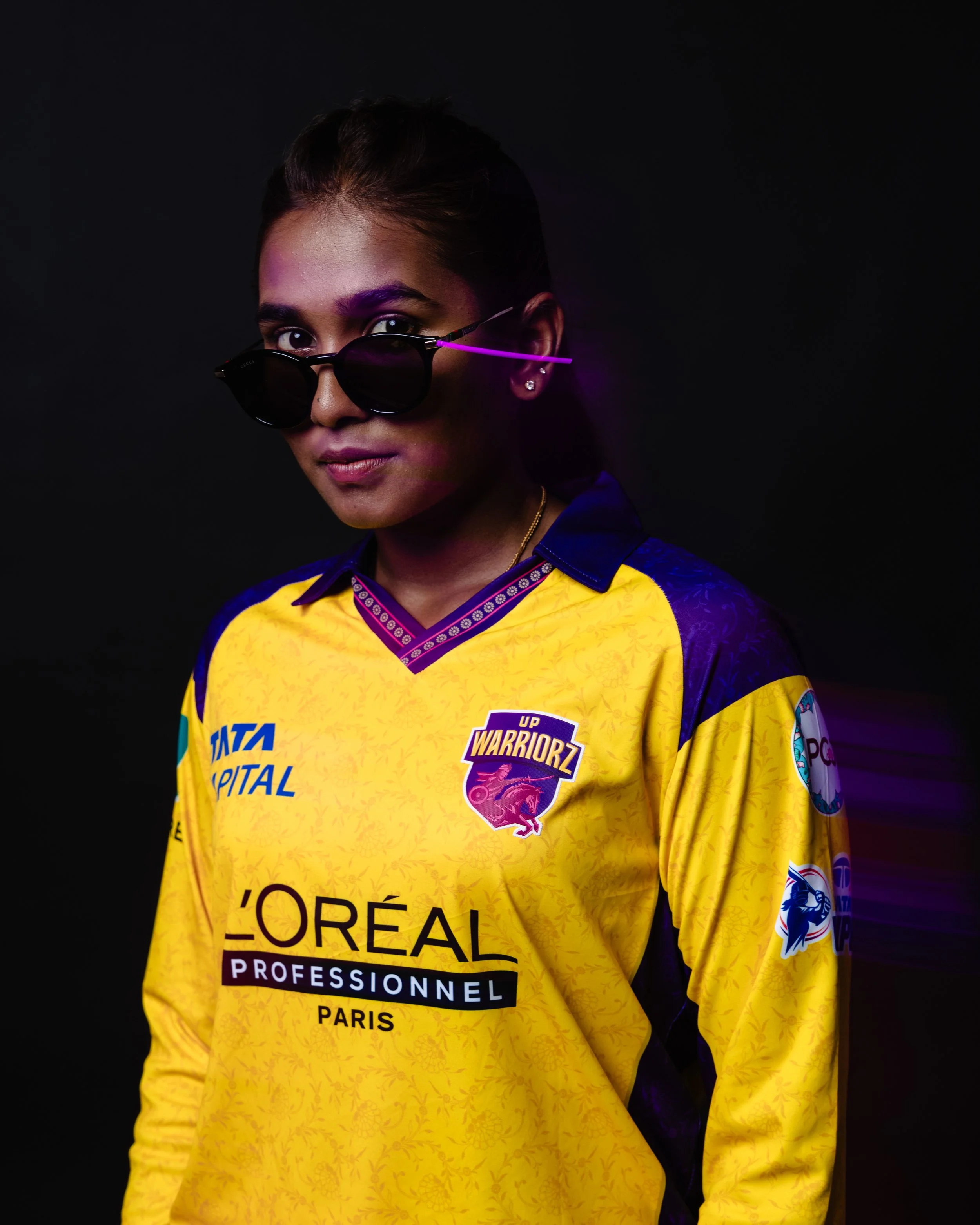A woman wearing sunglasses and a yellow sports jersey with various sponsor logos, including L'Oréal and Tata Capital, against a black background.