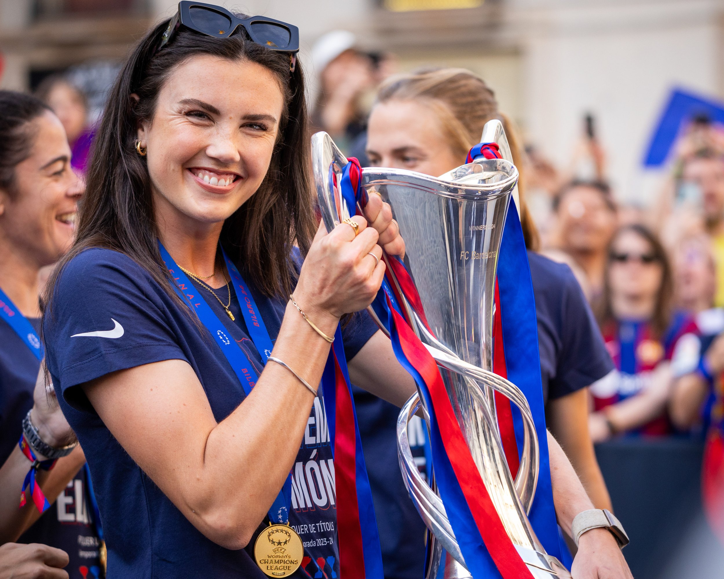 A smiling woman with long dark hair, wearing sunglasses on her head, holding a trophy cup with red and blue ribbons, surrounded by other women wearing medals and celebrating.
