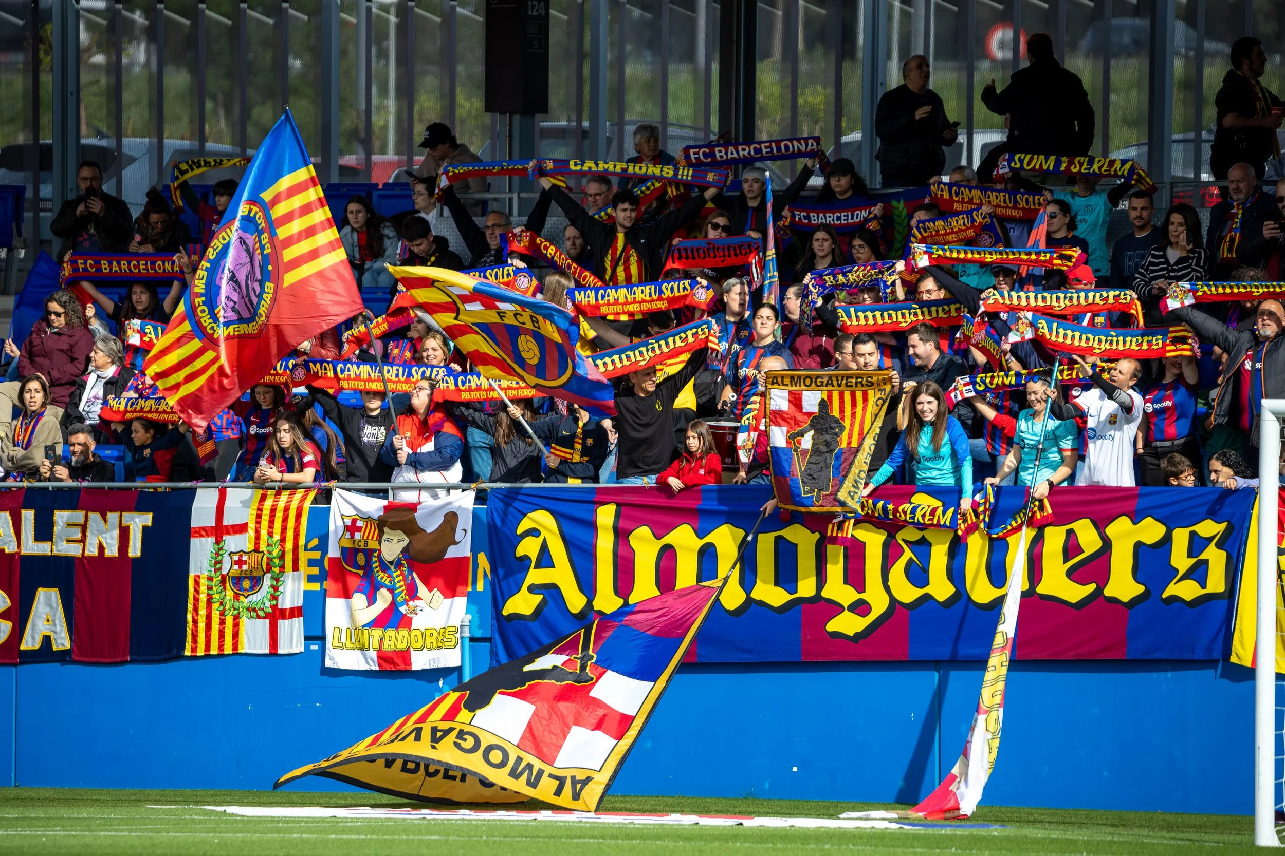 A group of football fans inside a stadium holding FC Barcelona scarves and flags, with some fans taking photos.