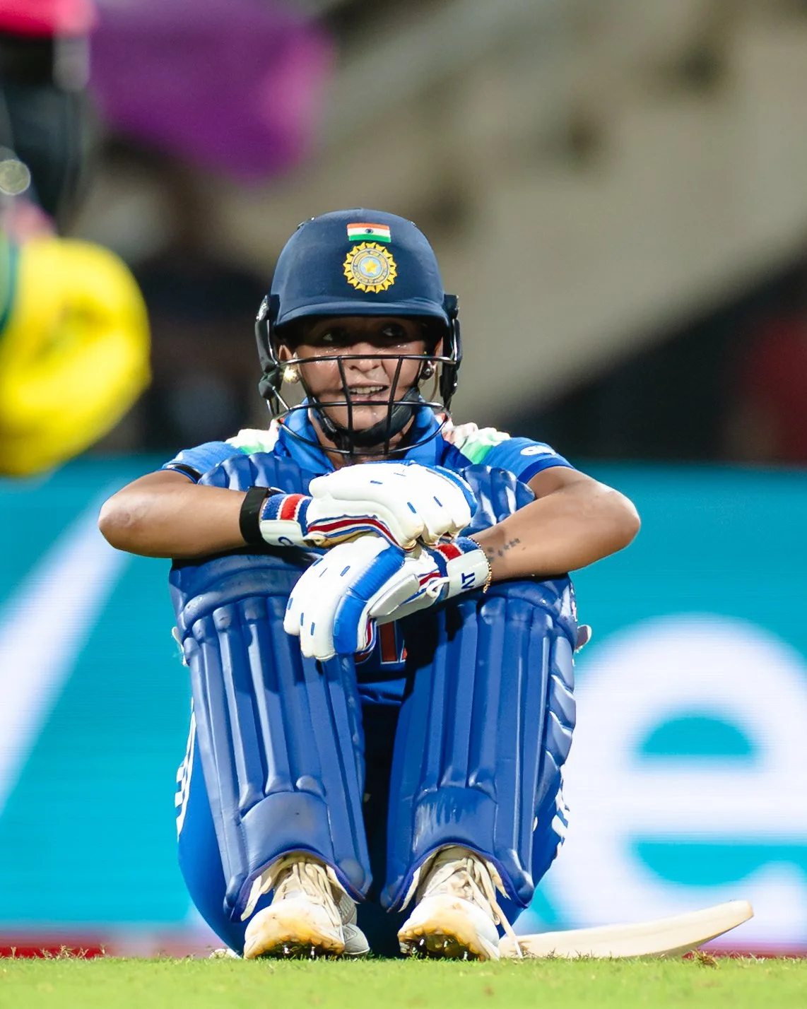 Indian female cricket player sitting down on the field in her full cricket gear, smiling, with her helmet and gloves on, during a match.