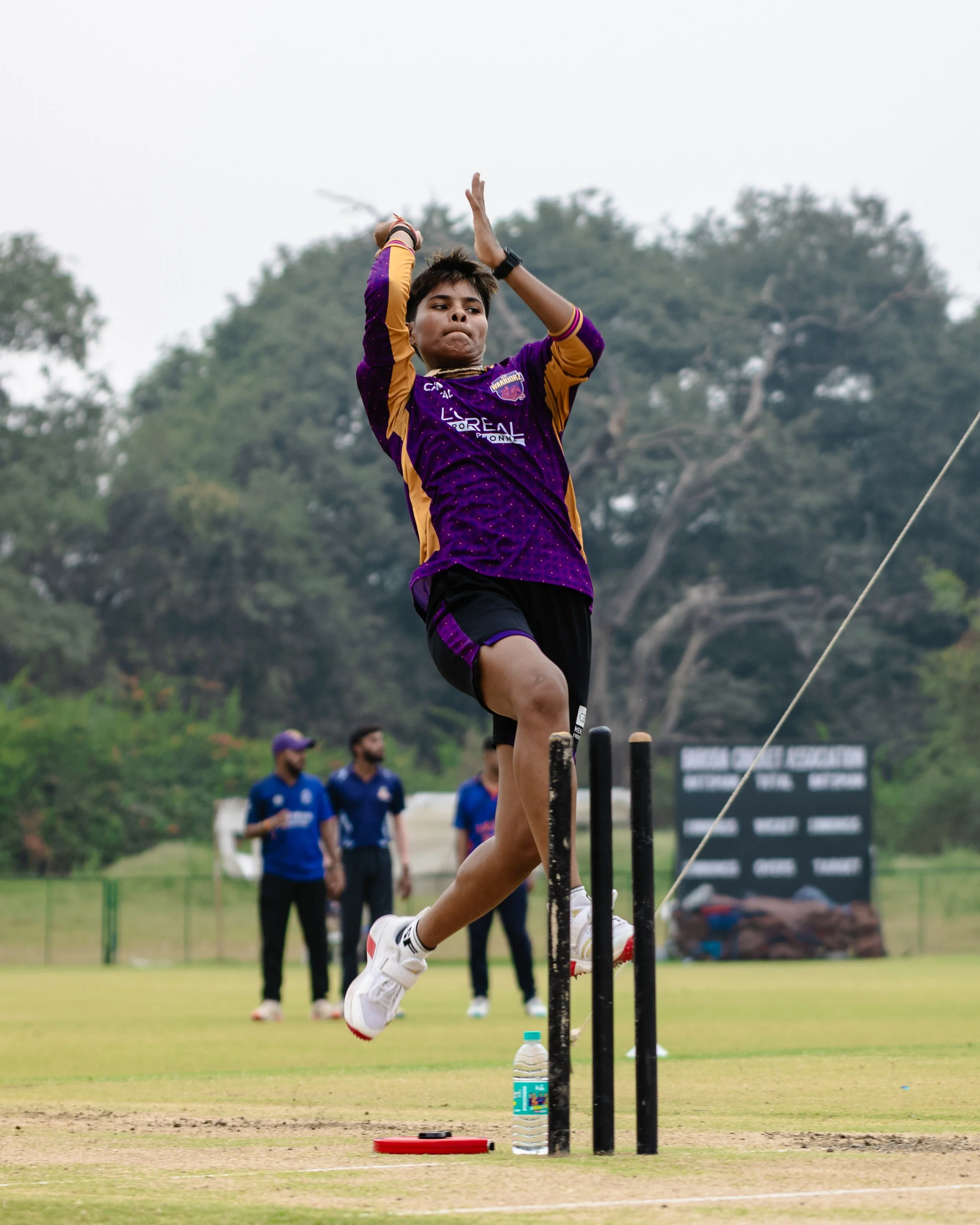 A person in a purple and yellow sports jersey is attempting to jump over hurdle on a sports field, with spectators and trees in the background.