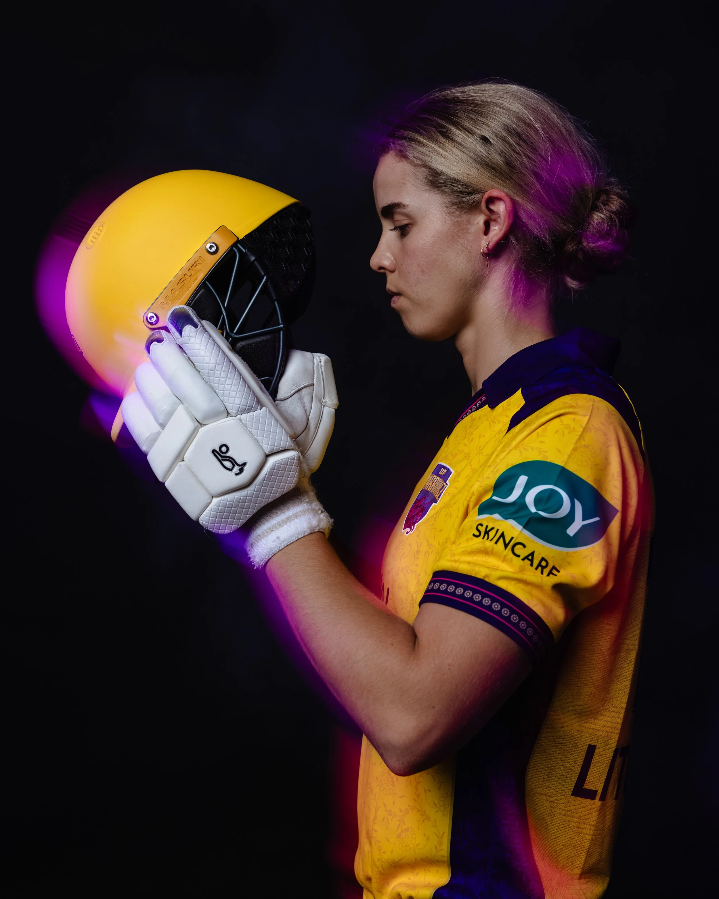 A female athlete dressed in a yellow sports jersey, holding a yellow helmet with a black face guard, touching foreheads with the helmet in front of her face against a black background.