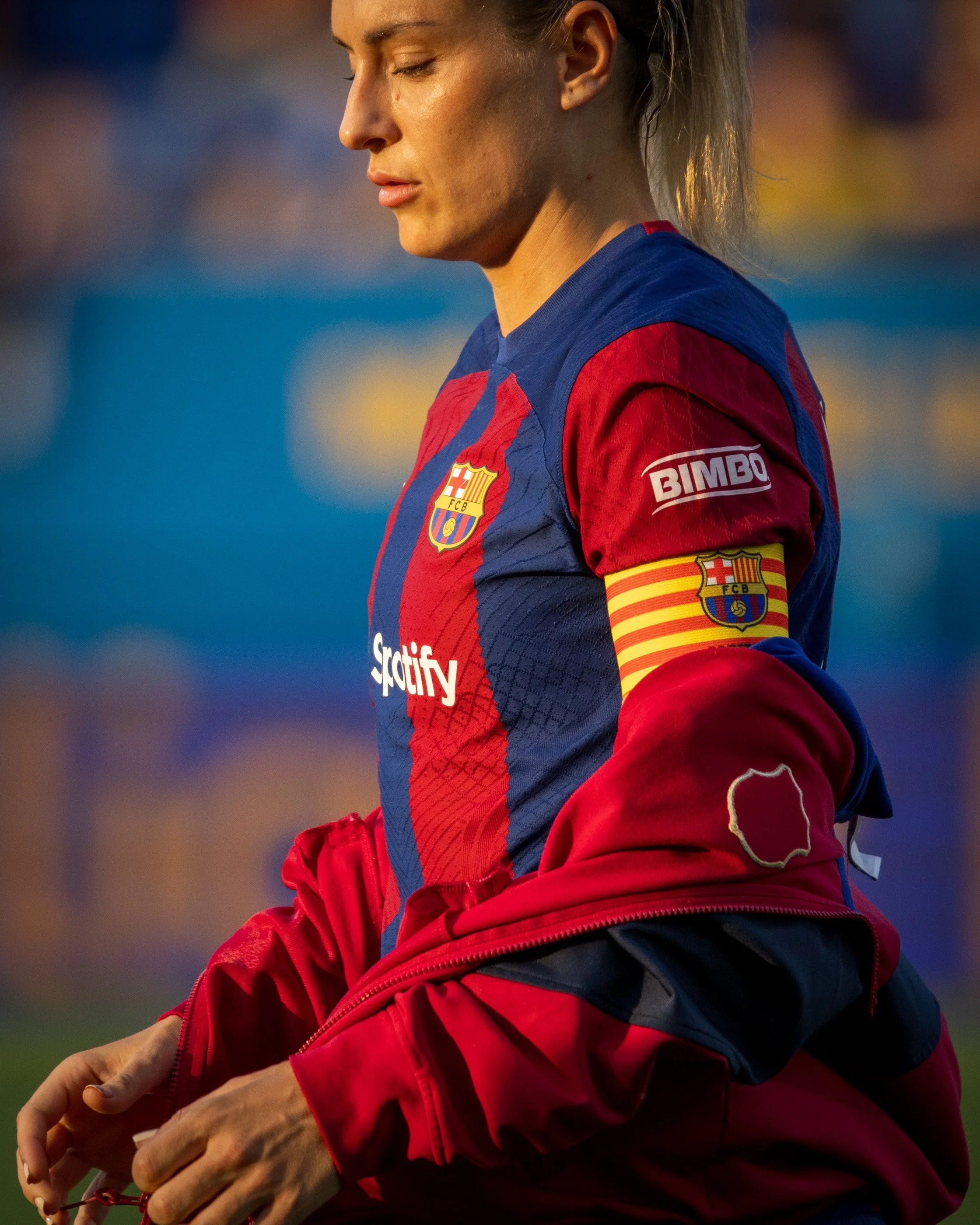 Close-up of a female athlete in a red and blue sports jersey with the FC Barcelona crest, adjusting her red jacket, with a focused expression and her eyes closed.