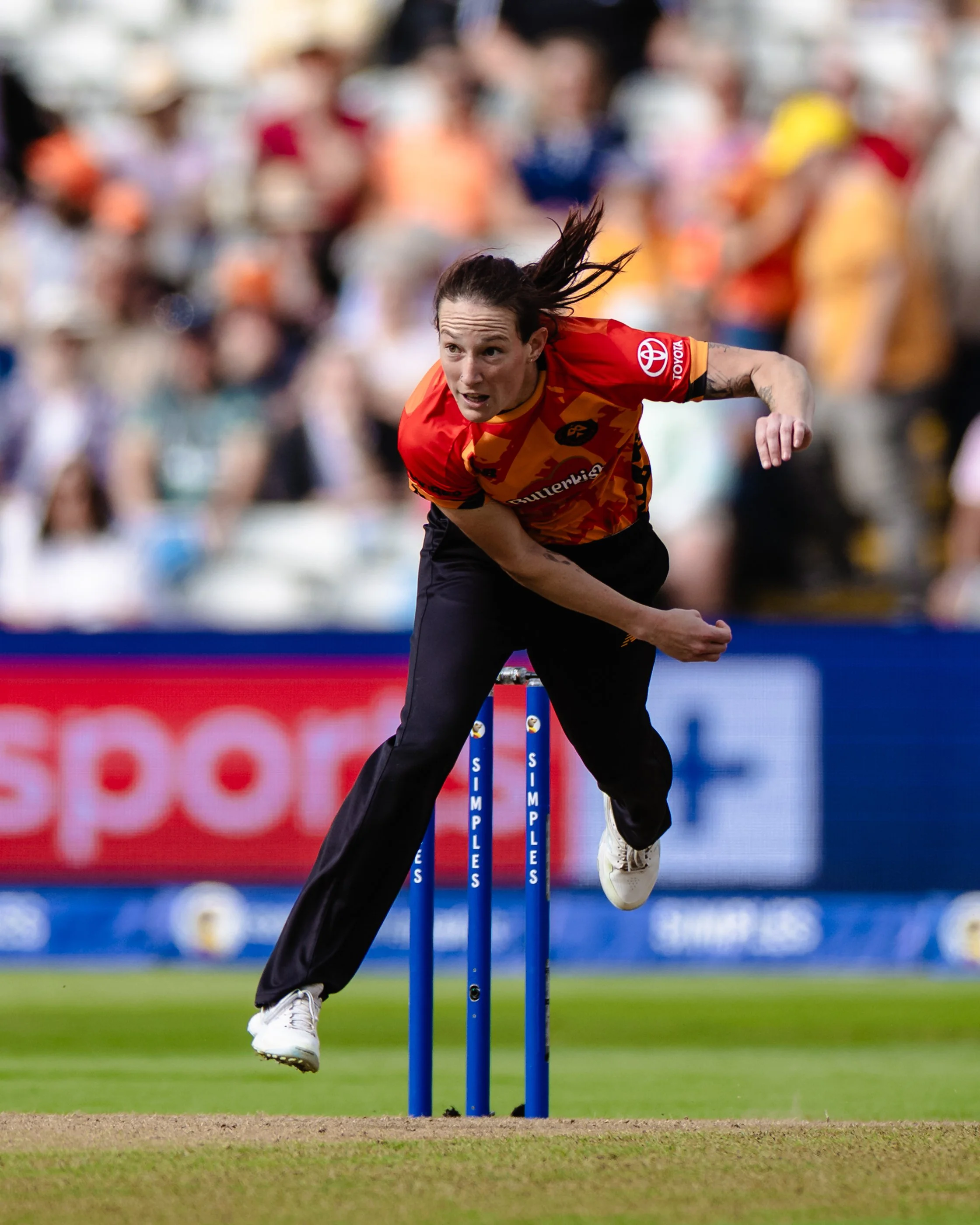 Female cricket player in red and orange jersey jumping to hit the ball with a cricket bat during a match.