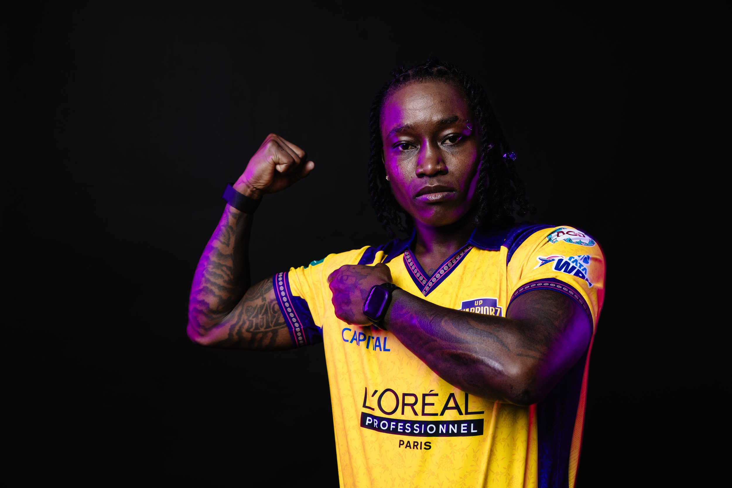A woman wearing a yellow sports jersey with sponsorship logos flexes her bicep while looking into the camera with a serious expression against a black background.