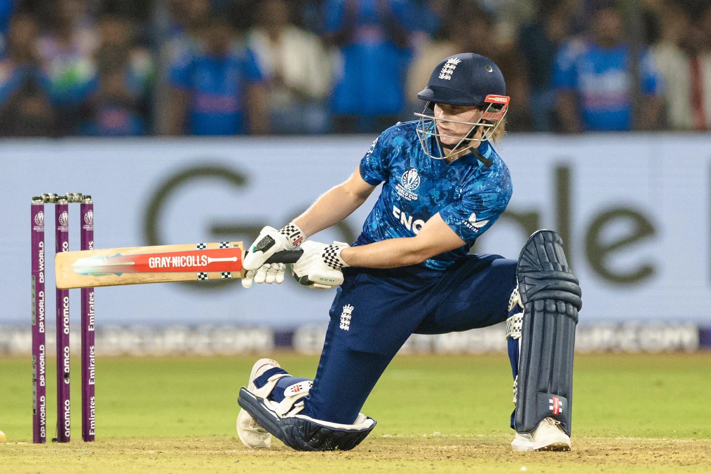 A female cricket player in blue uniform and helmet kneeling on the field, preparing to hit a cricket ball with her bat near the stumps with purple posts, during a match.