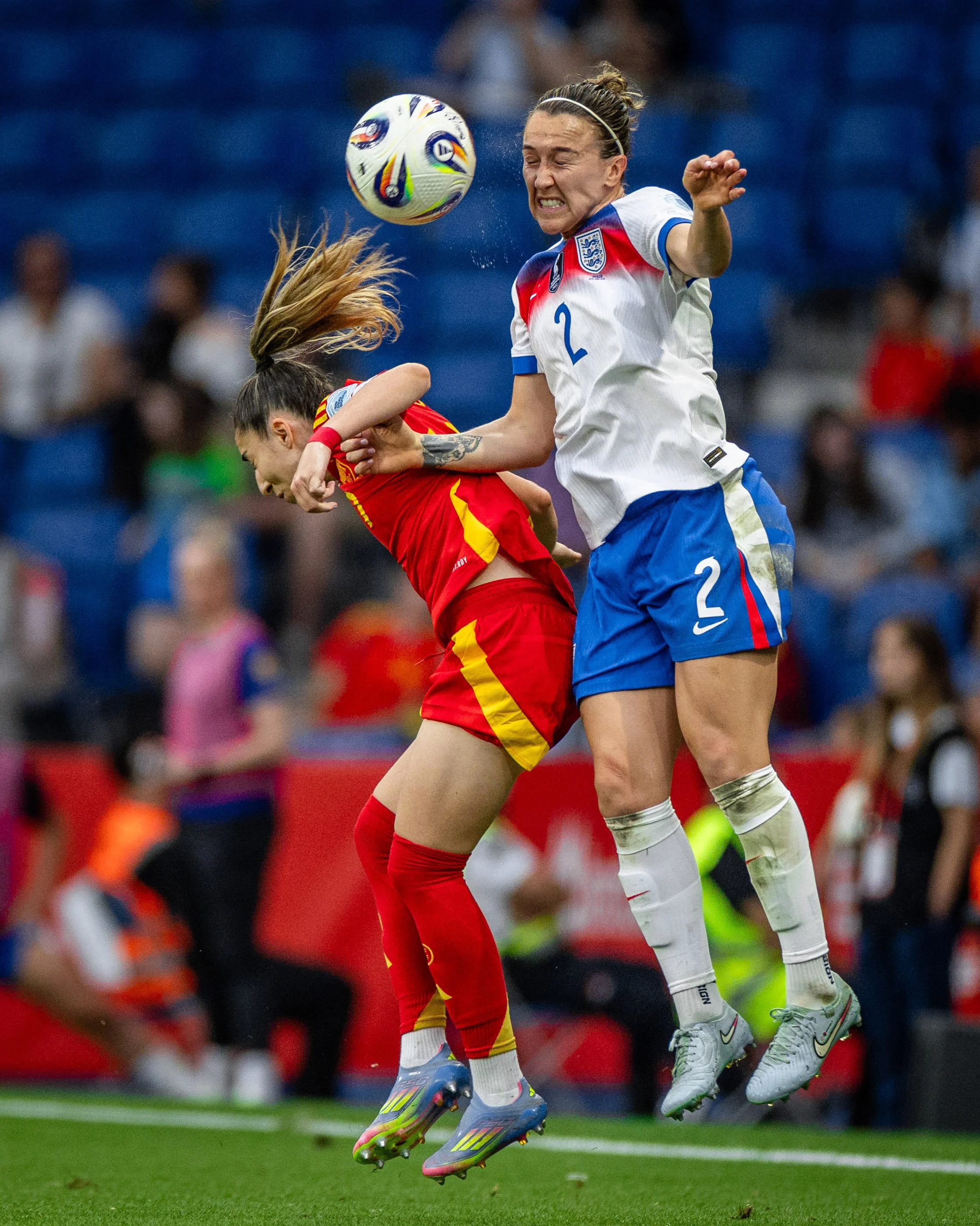 Two female soccer players are jumping and competing for the ball during a match. One player, wearing a white jersey with the number 2, appears to be heading the ball. The other player, wearing a red jersey with yellow accents, is also contesting for 