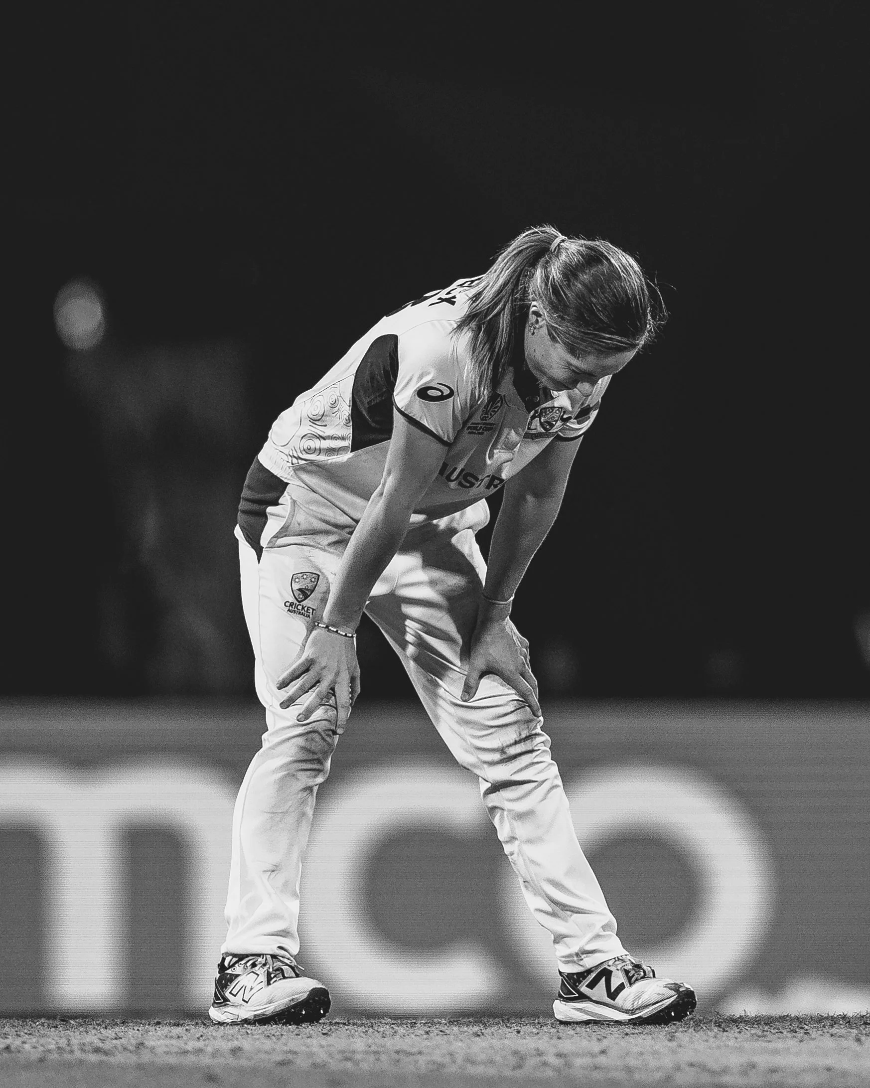 A female cricket player in uniform with a racket leaning forward on the ground, on a cricket field at night.
