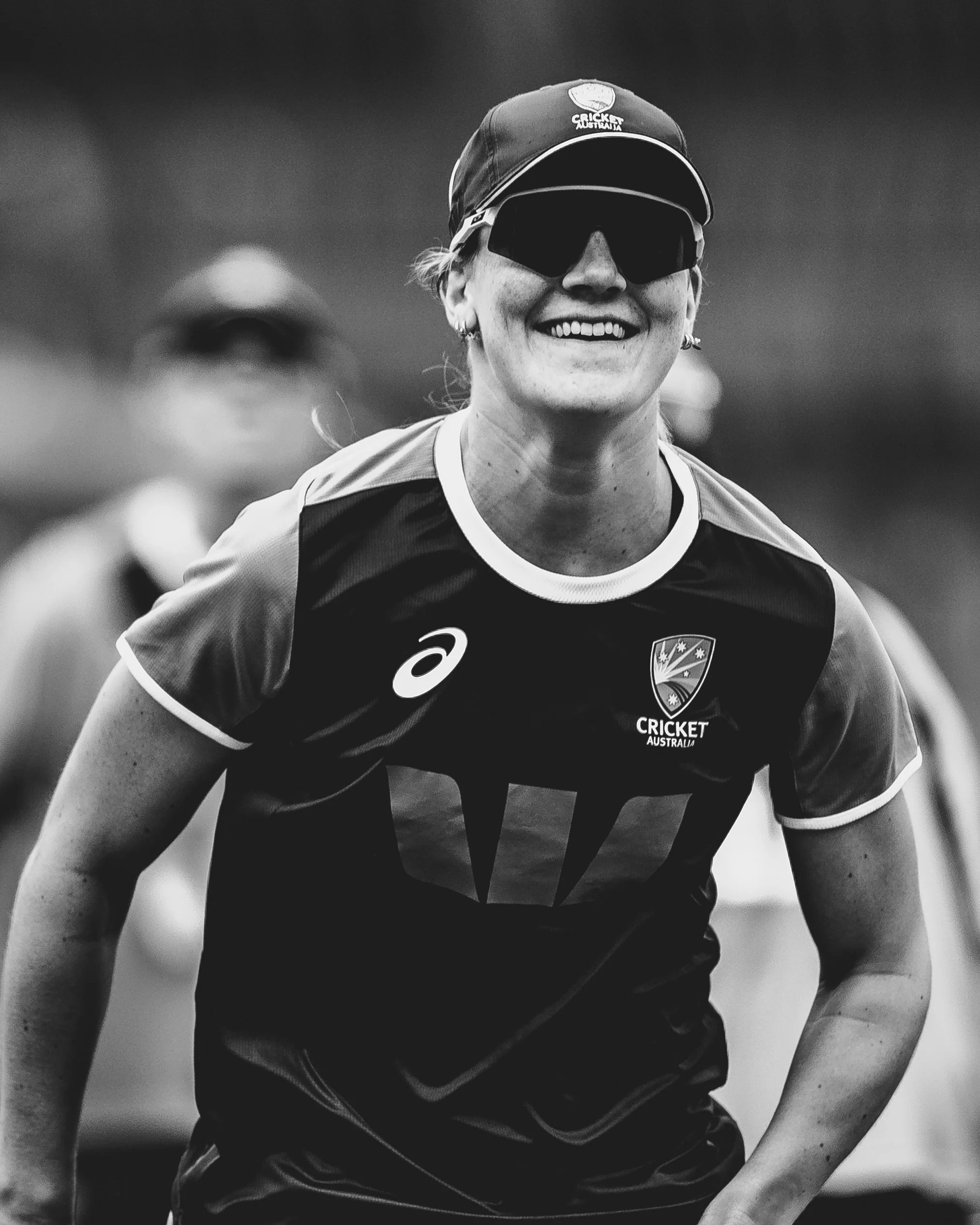 Black and white photo of a smiling female athlete wearing sunglasses, a cap, and a cricket jersey with logos.