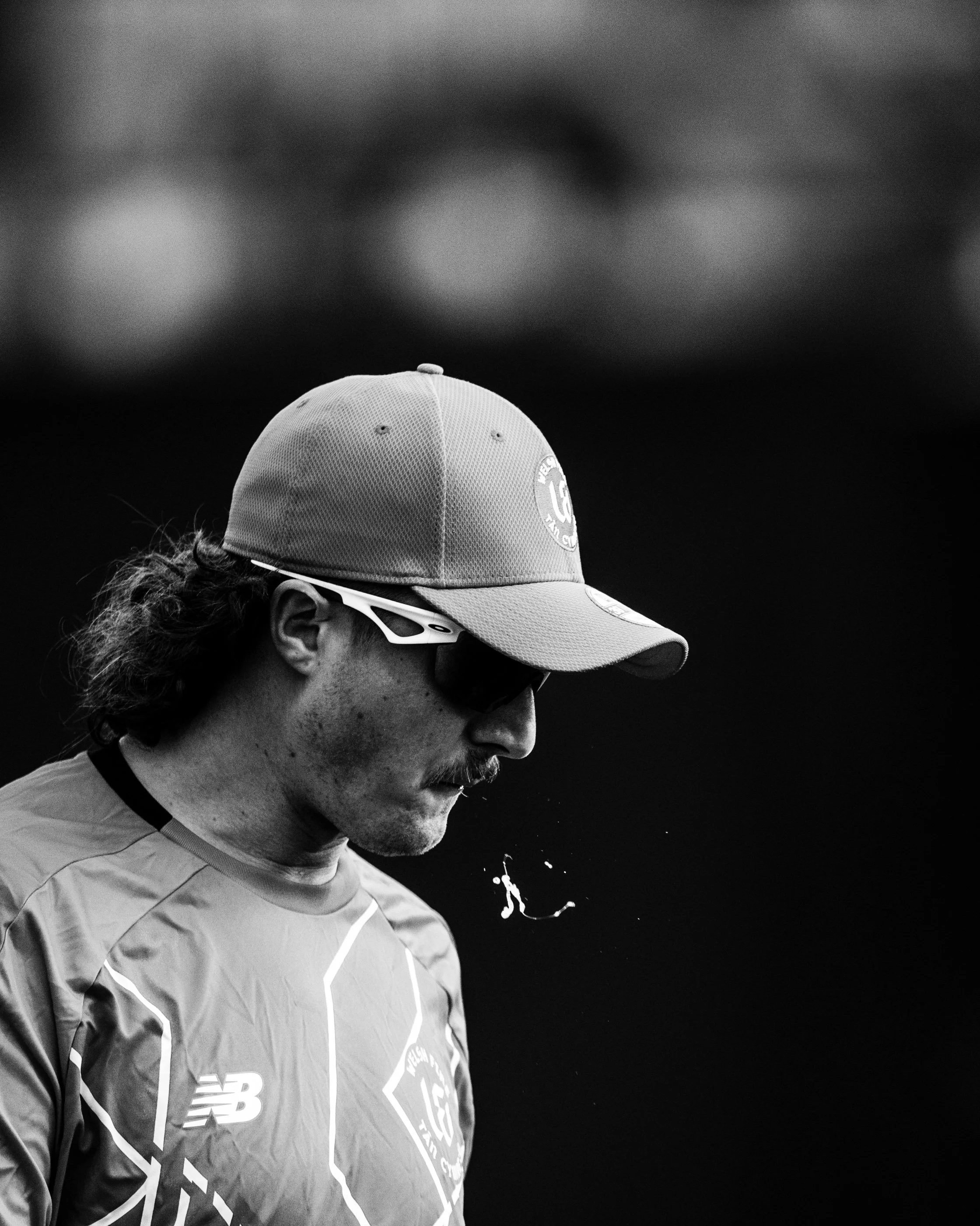 A person with curly hair wearing a baseball cap, sunglasses, and a sports jacket with logos, looking downward.