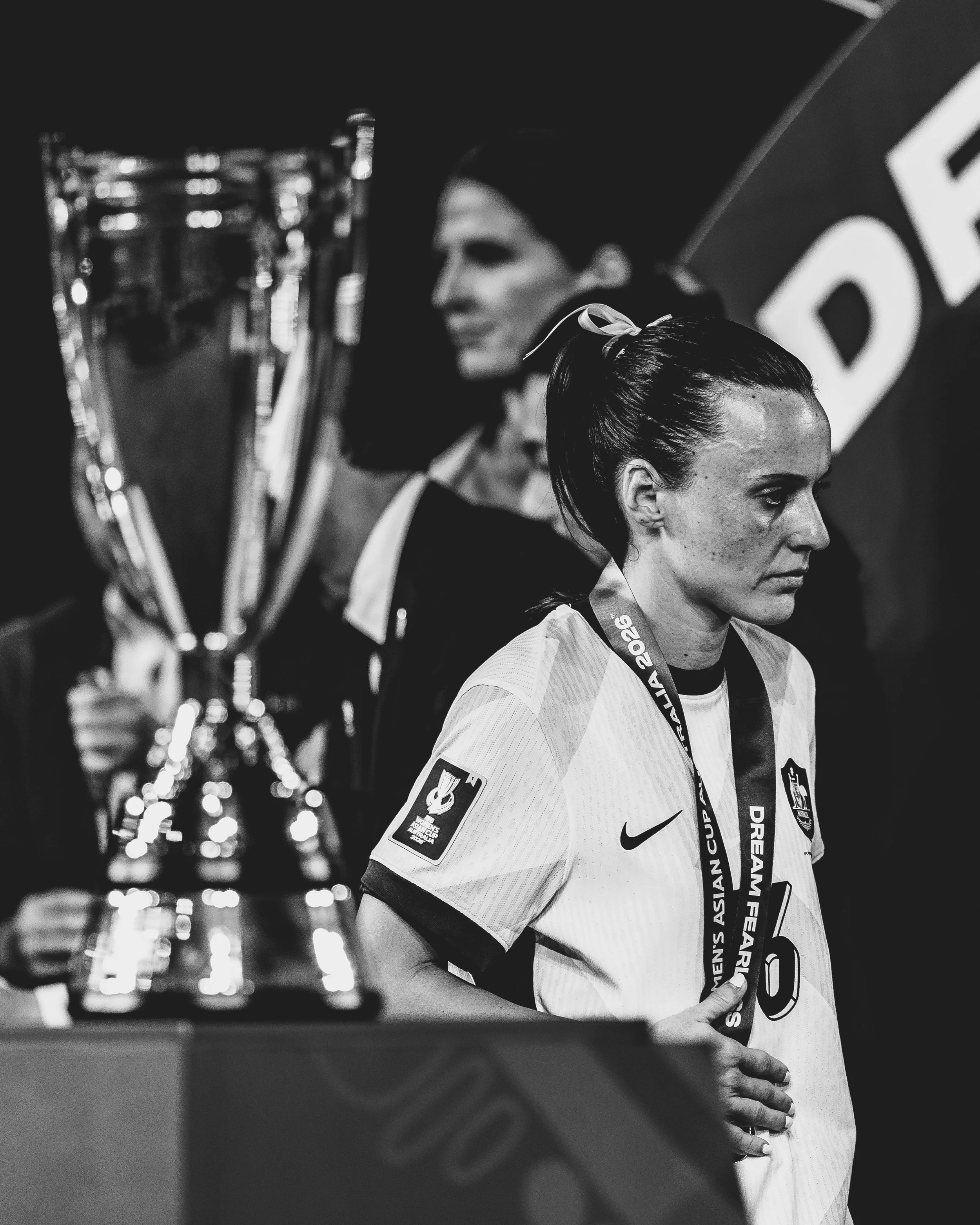 Two female soccer players standing side by side on a field, both wearing sports jerseys with medals around their necks. One is looking forward while the other looks to the side. Several trophies are in the foreground, slightly out of focus. The backg