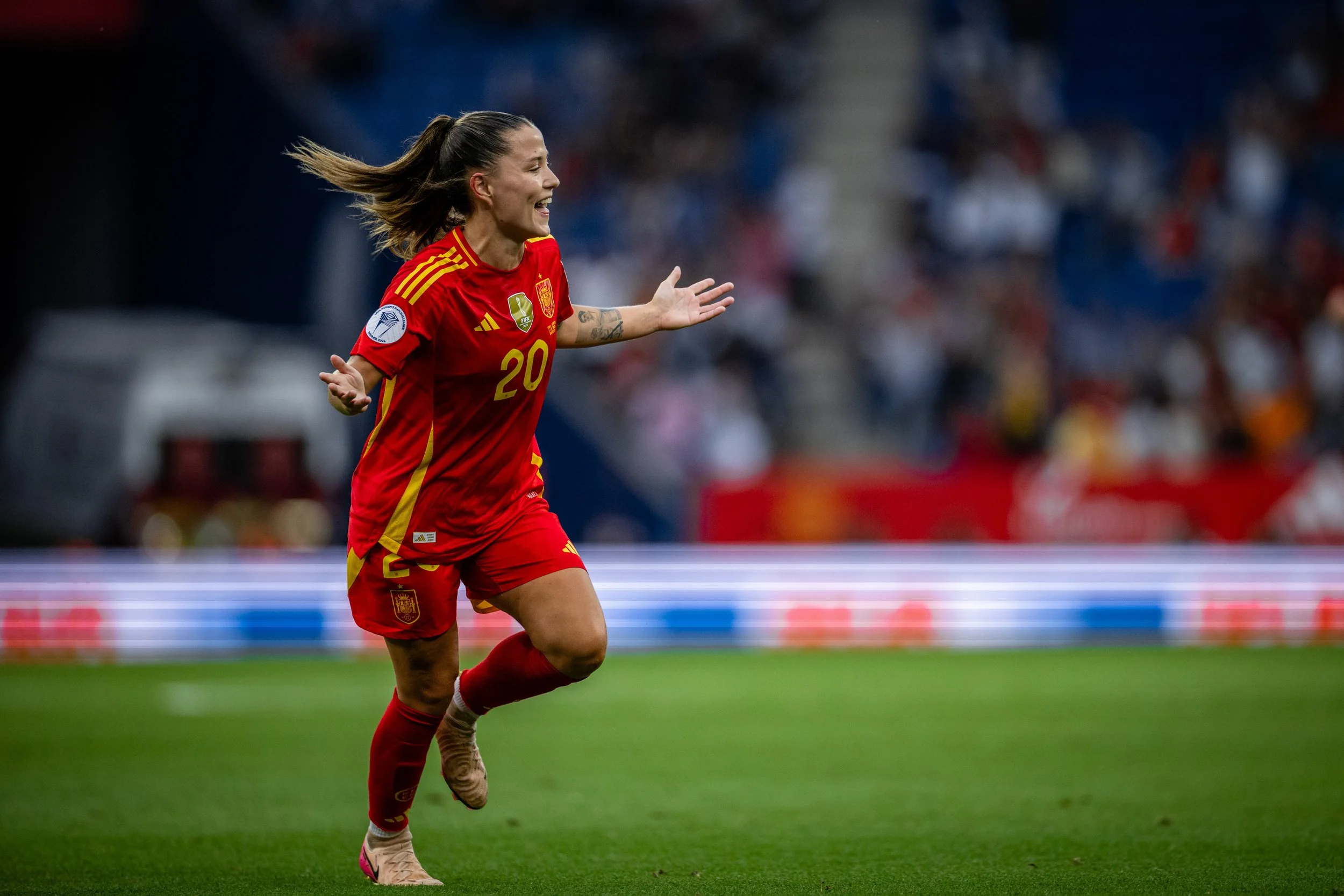 A female soccer player wearing a red jersey with yellow stripes and the number 20, running on a soccer field, celebrating.