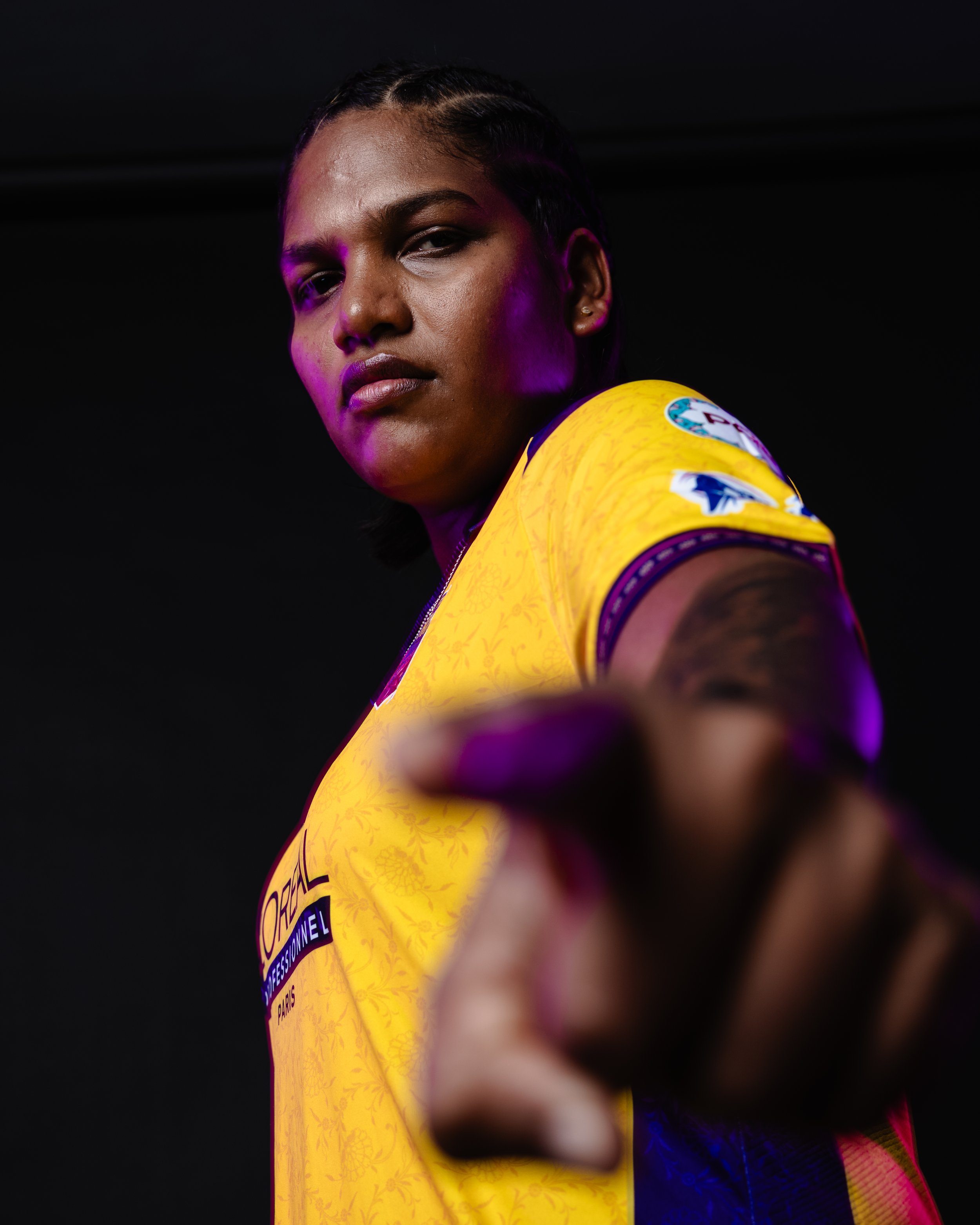 Close-up of a female athlete wearing a yellow sports jersey with patches, looking confidently at the camera, with purple lighting effects and a dark background.
