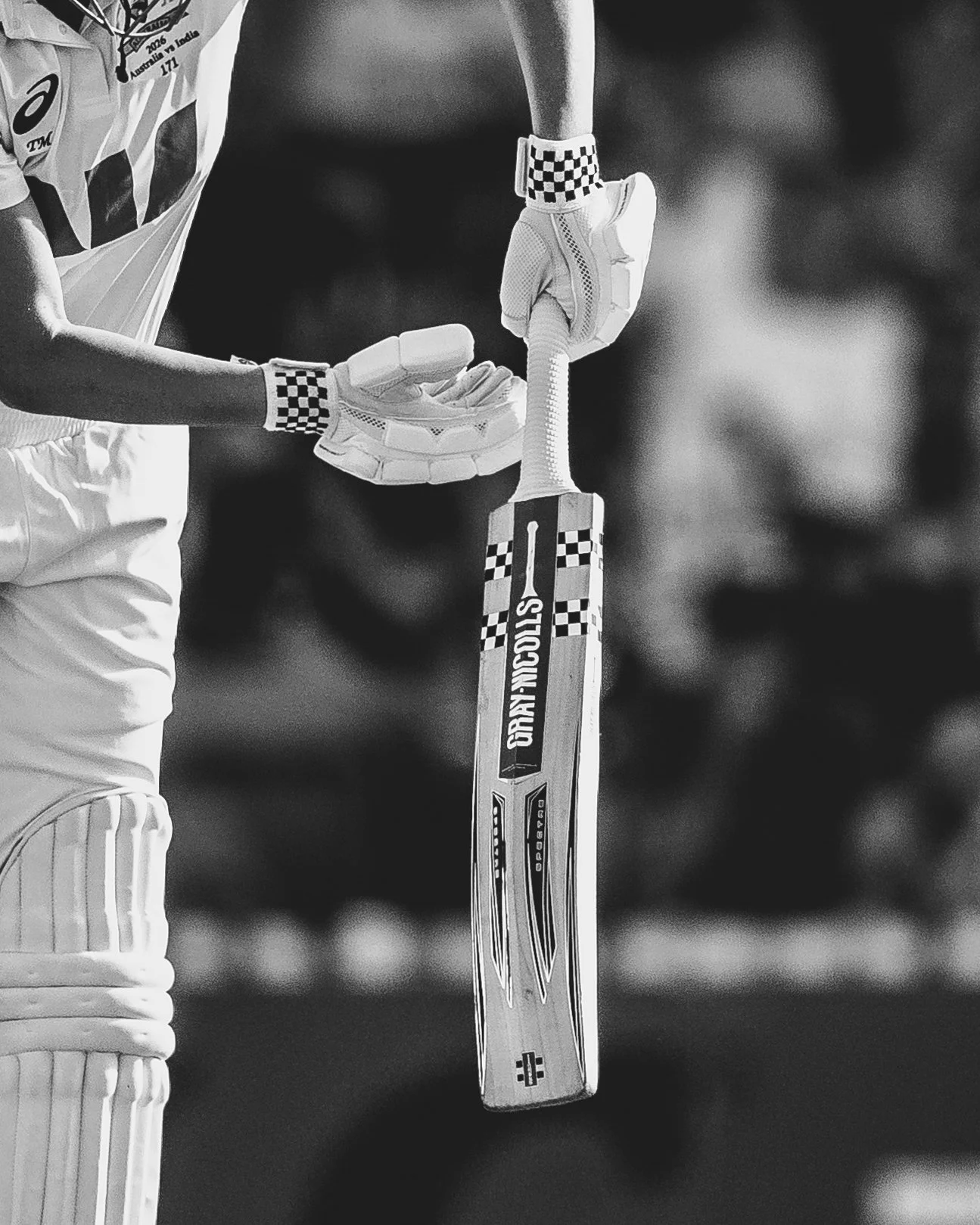 Close-up of a cricket player's gloved hand holding a cricket bat with a checkered pattern on the wristband, and a cricket ball resting on the bat's blade.