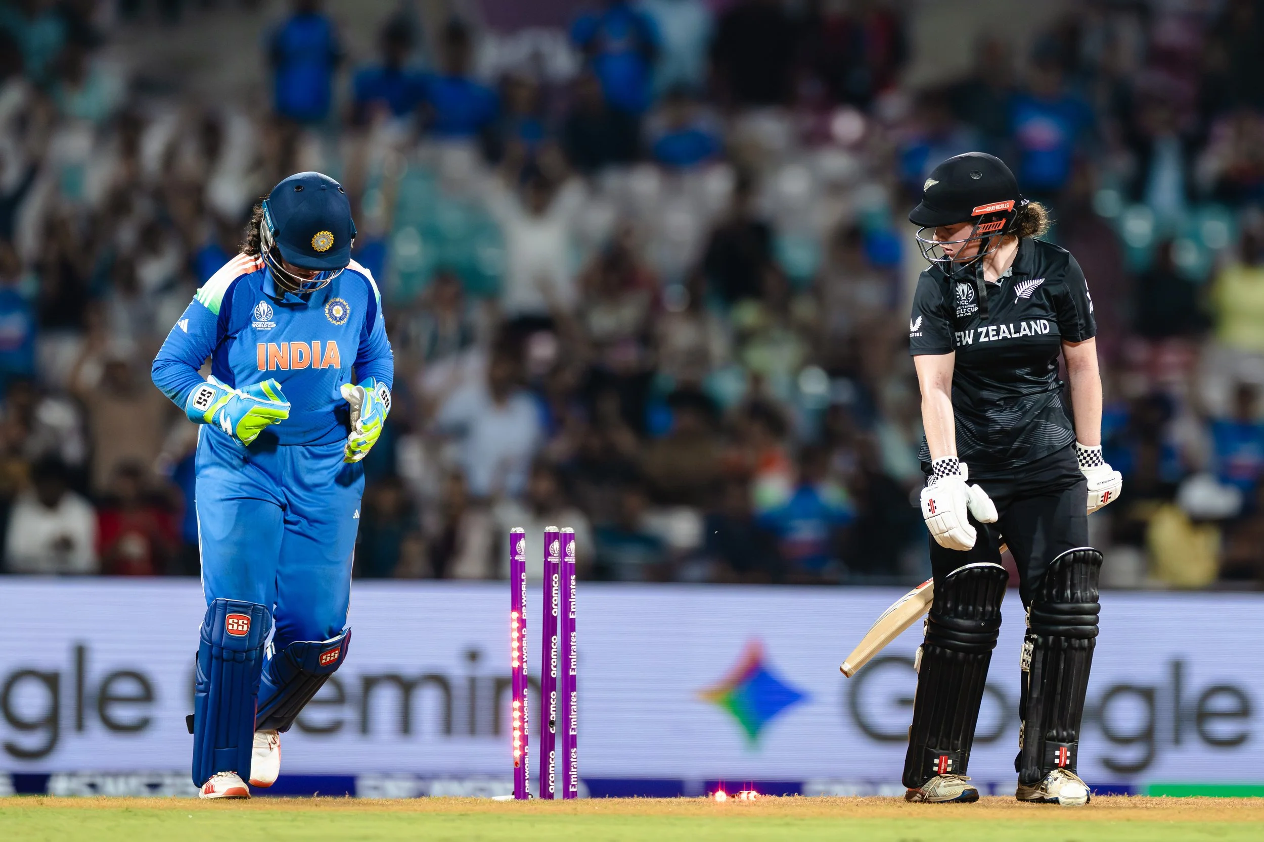 Cricketers from India and New Zealand on the cricket field during a match, with one team in blue uniform and another in black, at night with a blurred crowd in the stands.