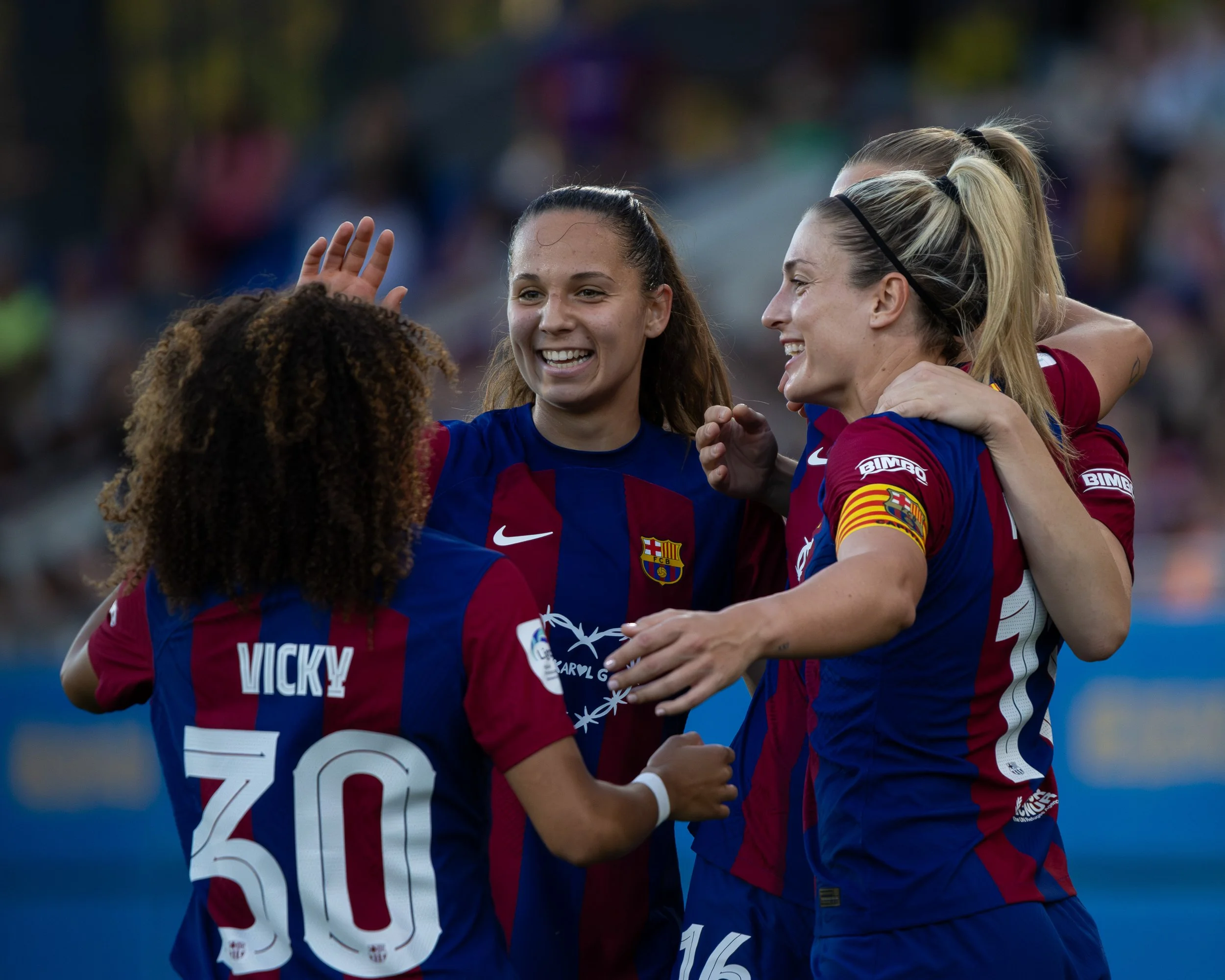 A group of female soccer players wearing red and blue jerseys celebrating on the field, with one woman smiling and others hugging.