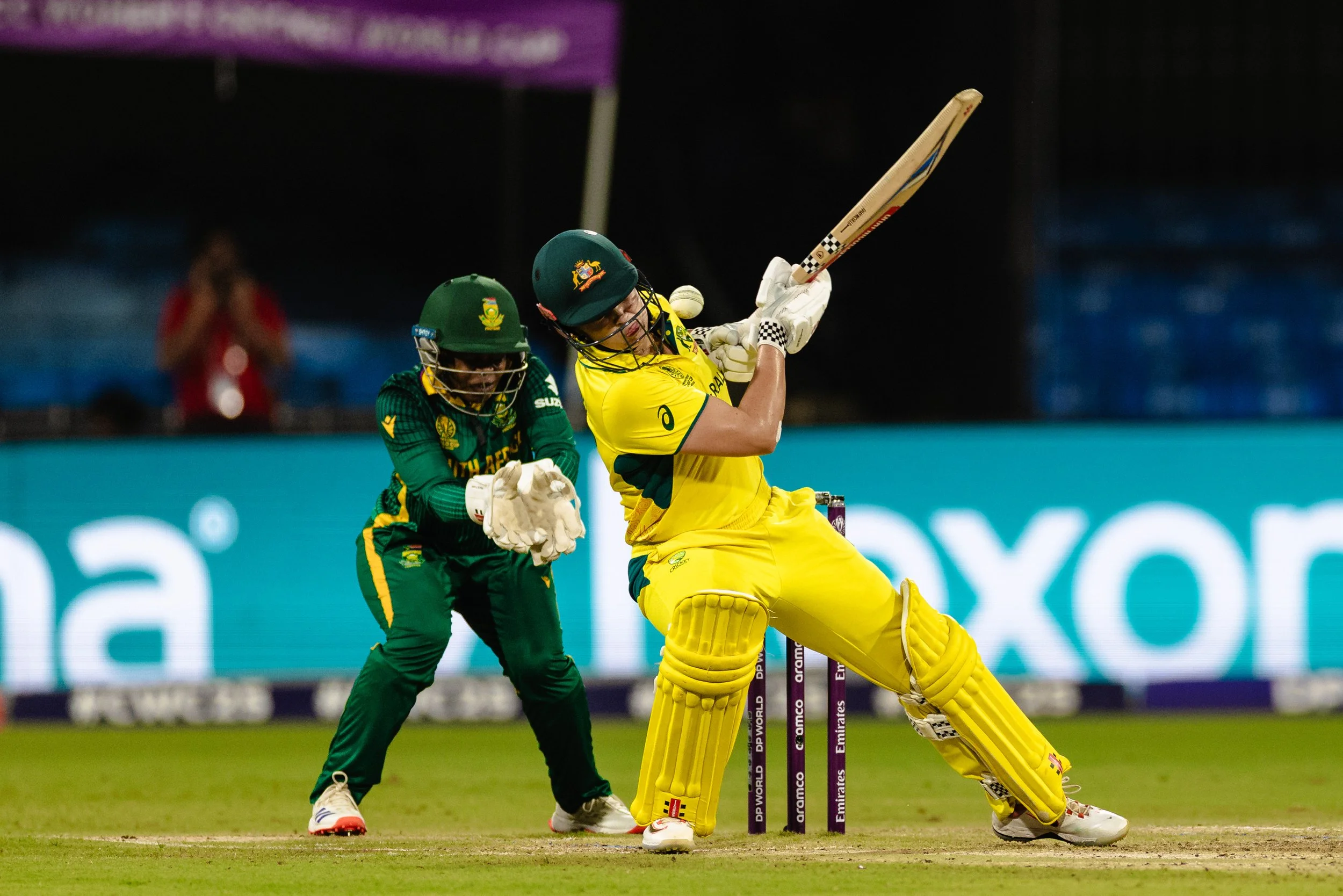 A cricket player in yellow uniform batting, with a wicket keeper in green uniform behind him, during a match at night.
