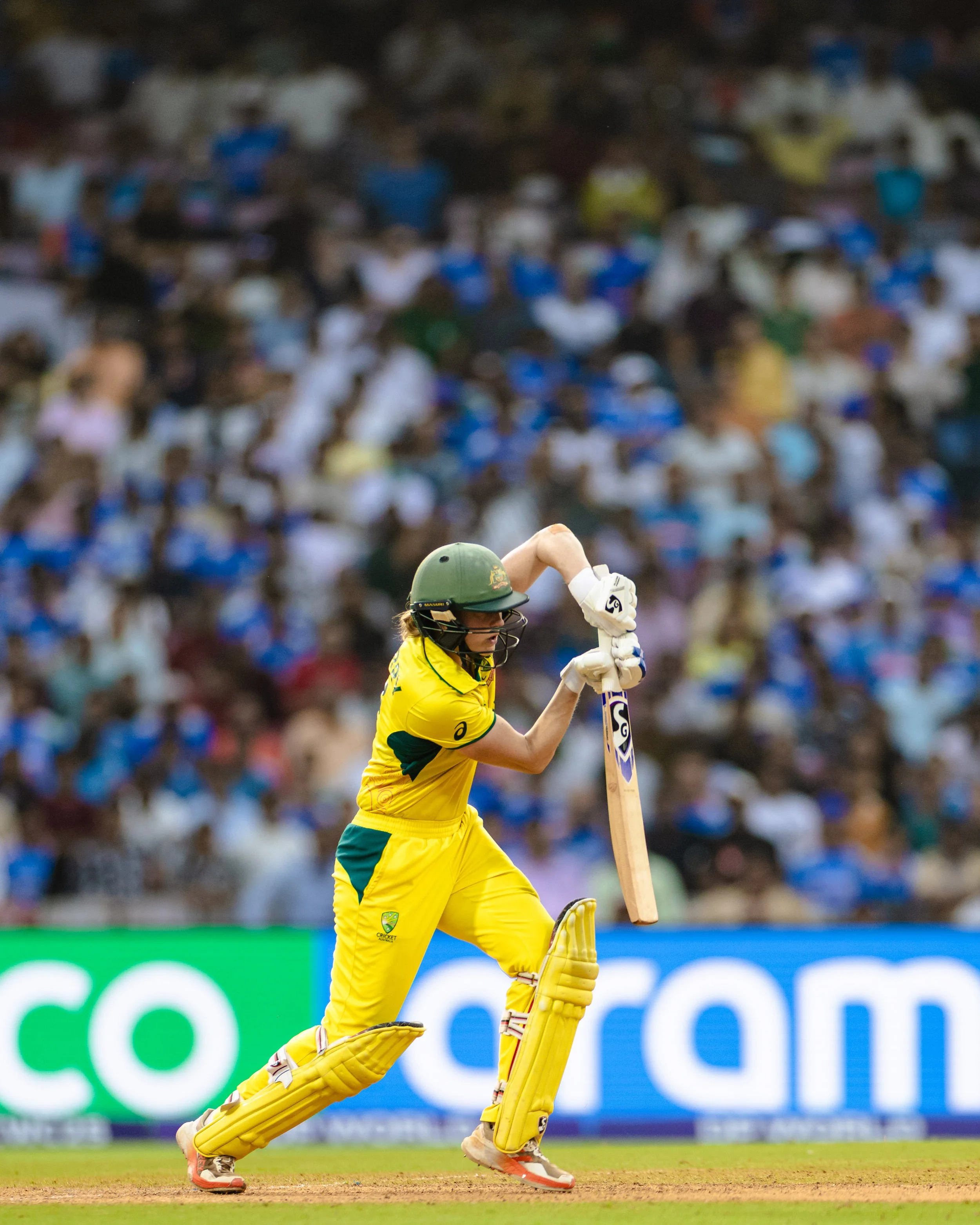 A cricket player in yellow uniform and helmet strikes a ball during a match with a large crowd in the background.