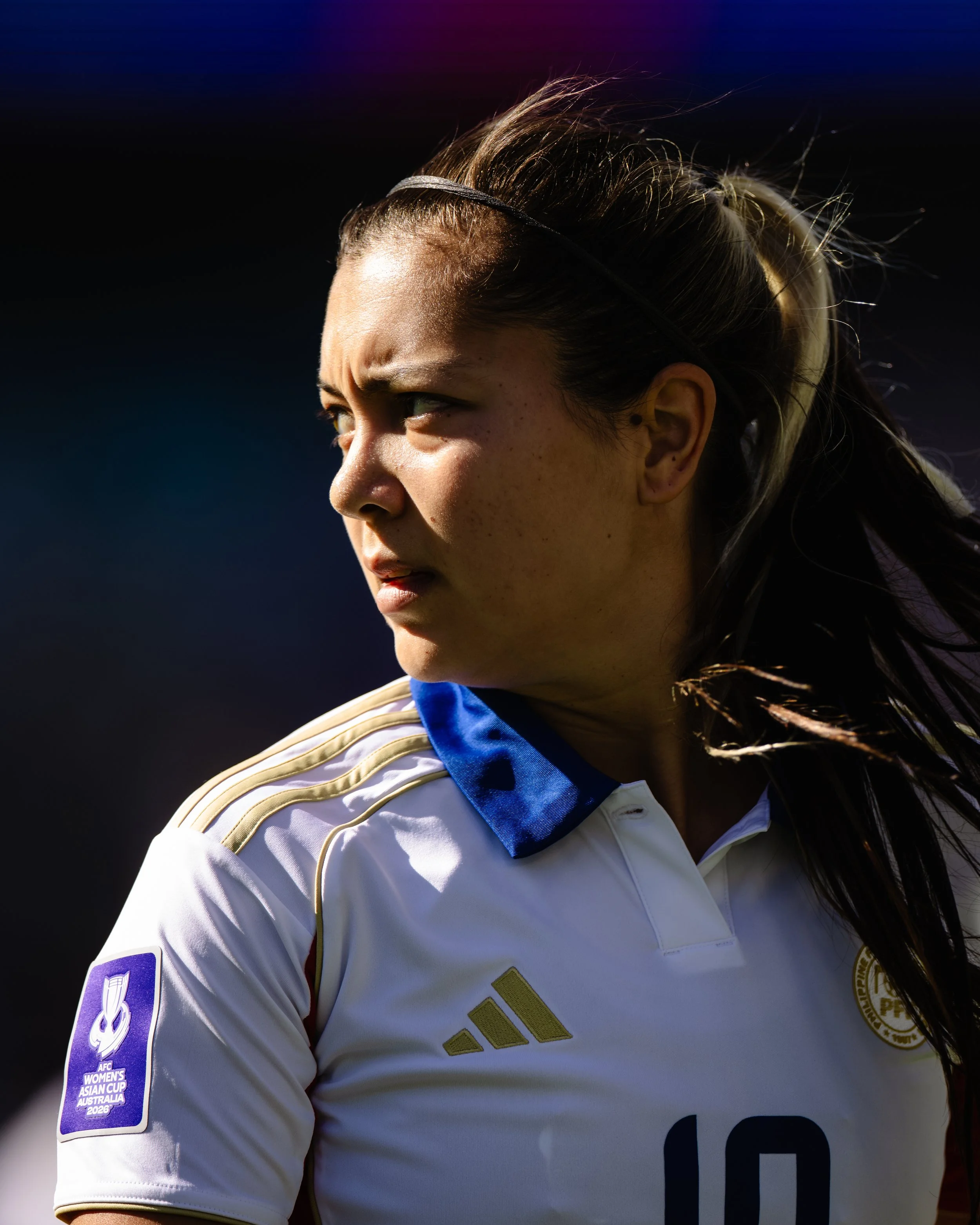Close-up of a female athlete wearing a white sports jersey with gold and blue accents, featuring a patch for the AFC Women's Asian Cup Australia 2026 on her sleeve and an emblem on her chest, with a focused expression looking to the side.