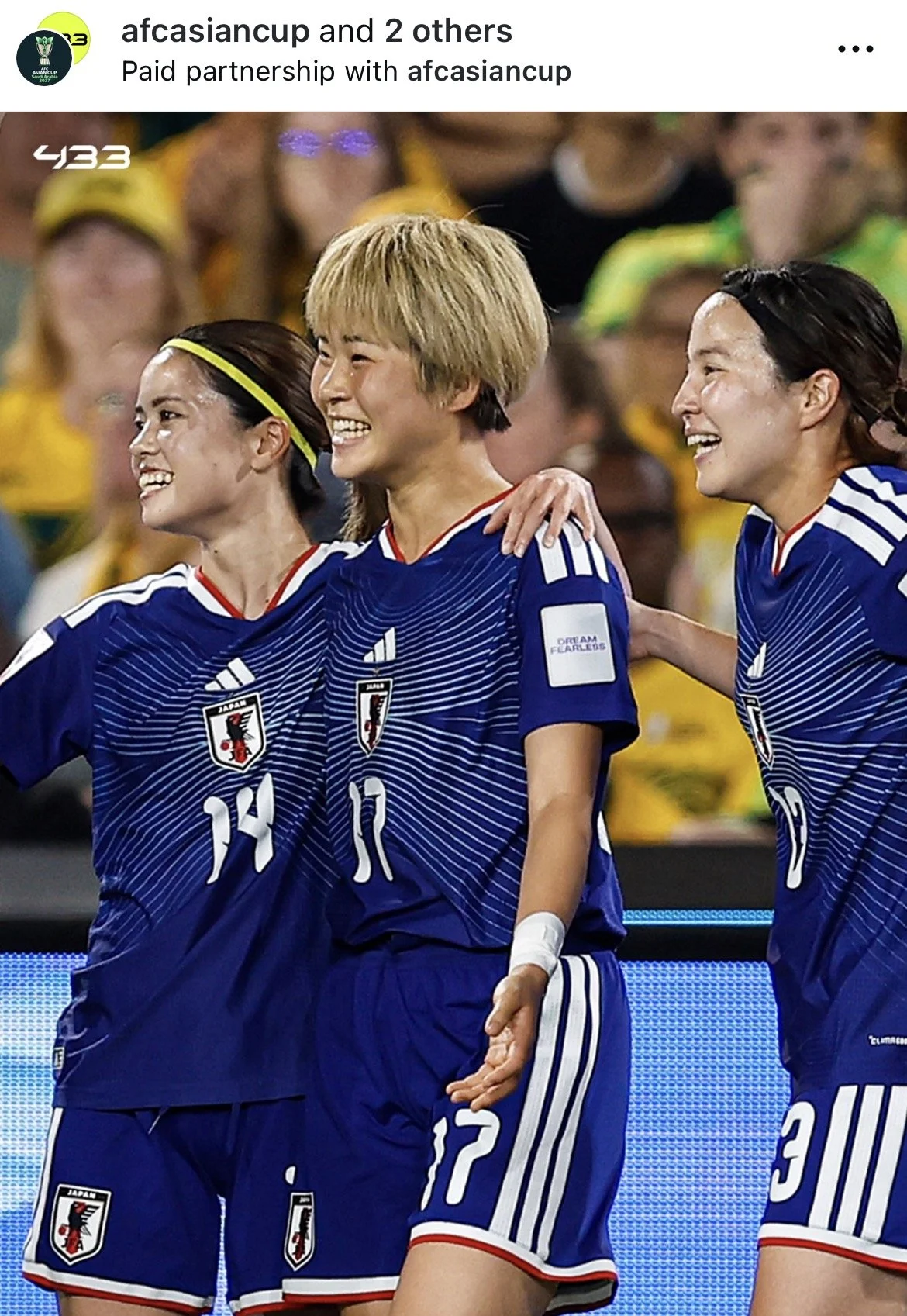 Three female soccer players from Japan in blue jerseys celebrating together on the field.