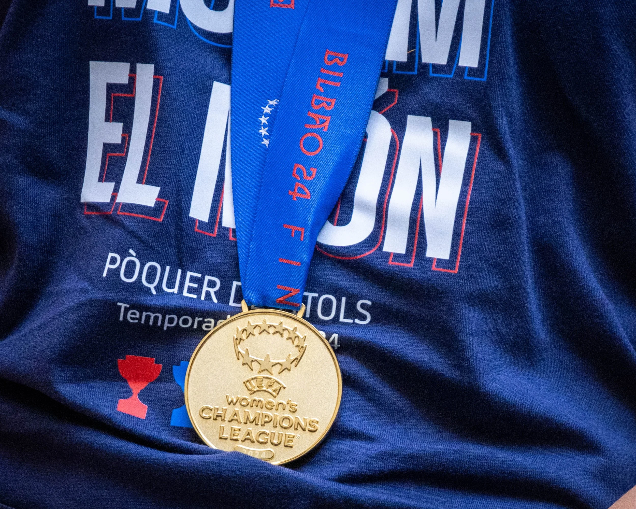 Close-up of a gold medal hanging from a blue ribbon with red text, placed on a dark blue fabric with printed text and graphics related to a sports event.