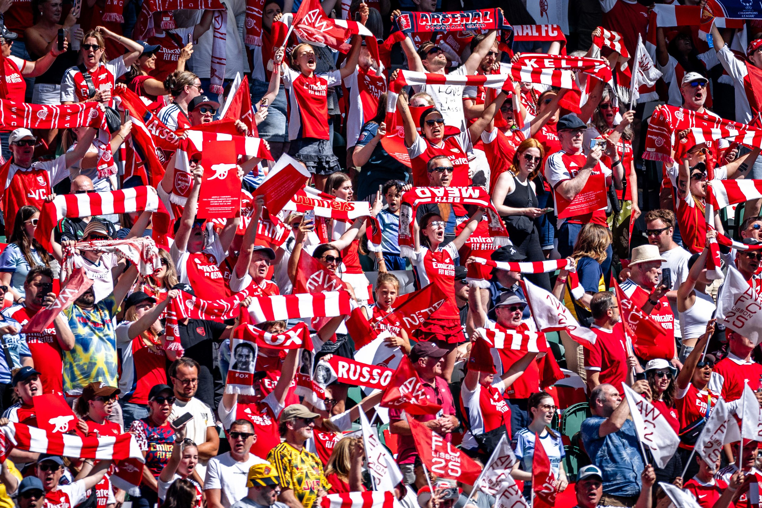 Crowd of enthusiastic soccer fans dressed in red and white, holding scarves and flags, cheering at a stadium event.