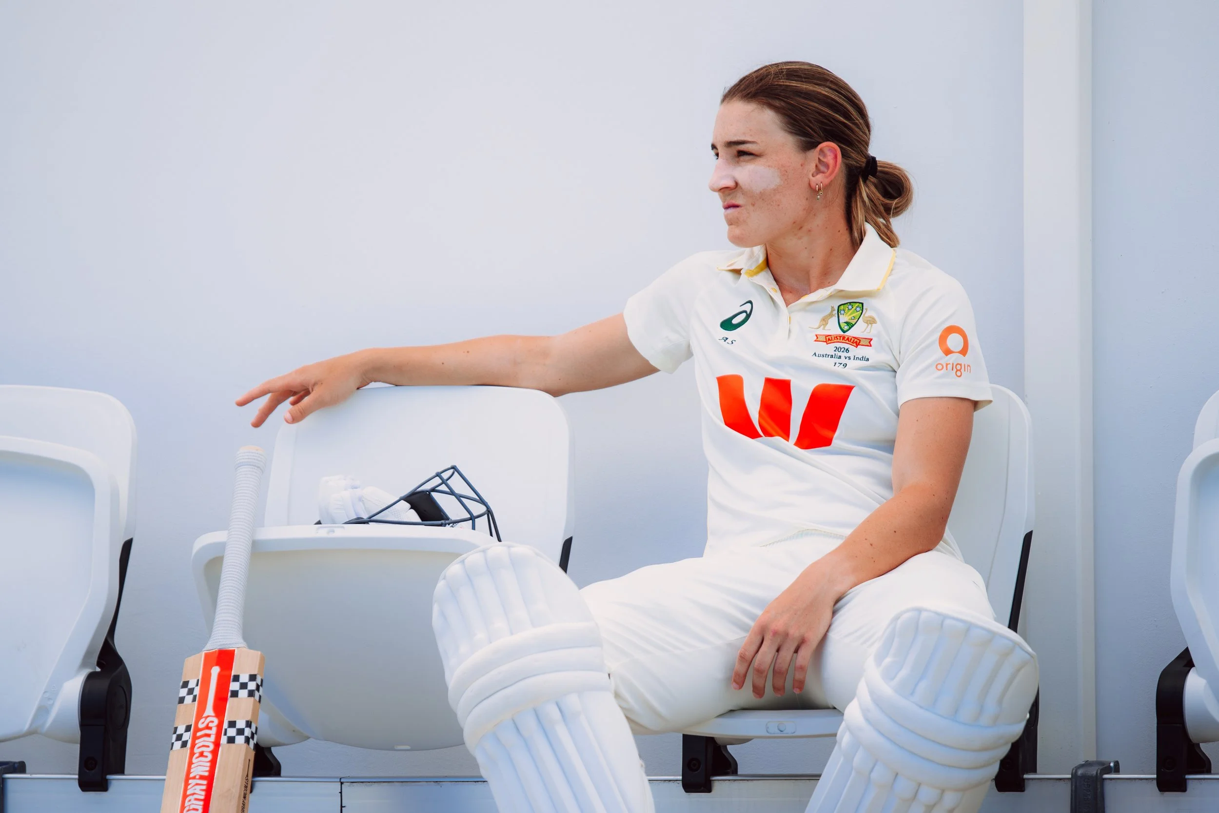 A female athlete with brown hair tied back, wearing a white sports uniform with logos, seated on a white chair, looking to her left with a focused expression. She is surrounded by white seats and sports equipment.