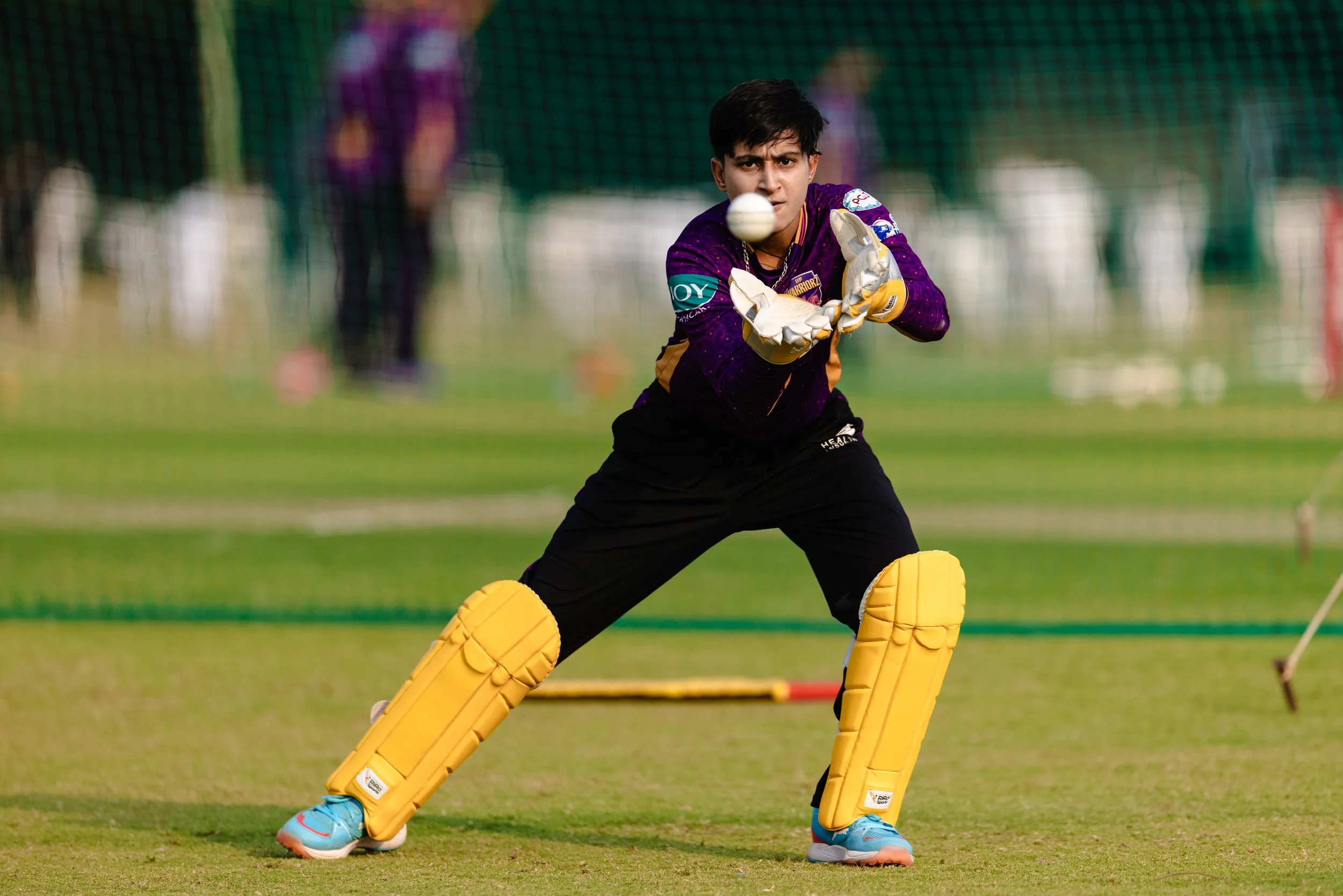 A young male cricket player in purple and yellow uniform with protective gear, including yellow leg pads and gloves, is in a ready stance preparing to catch a cricket ball on a field.