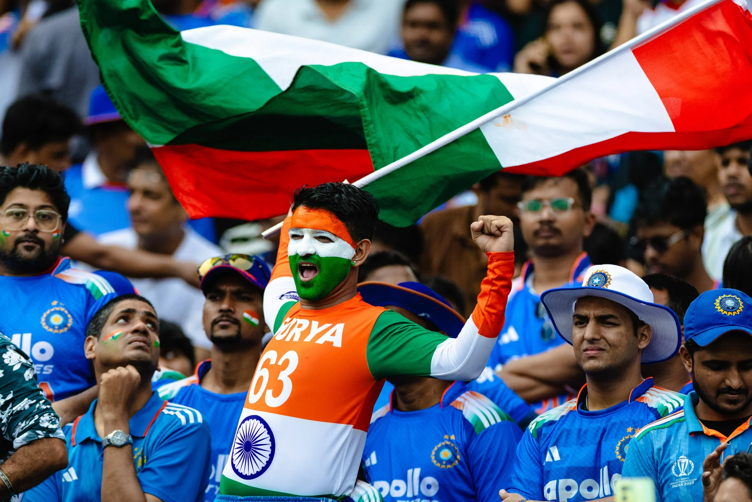 A crowd of cricket fans celebrating, with an Indian cricket player holding the Indian flag. Many spectators are dressed in blue jerseys, some with face paint.