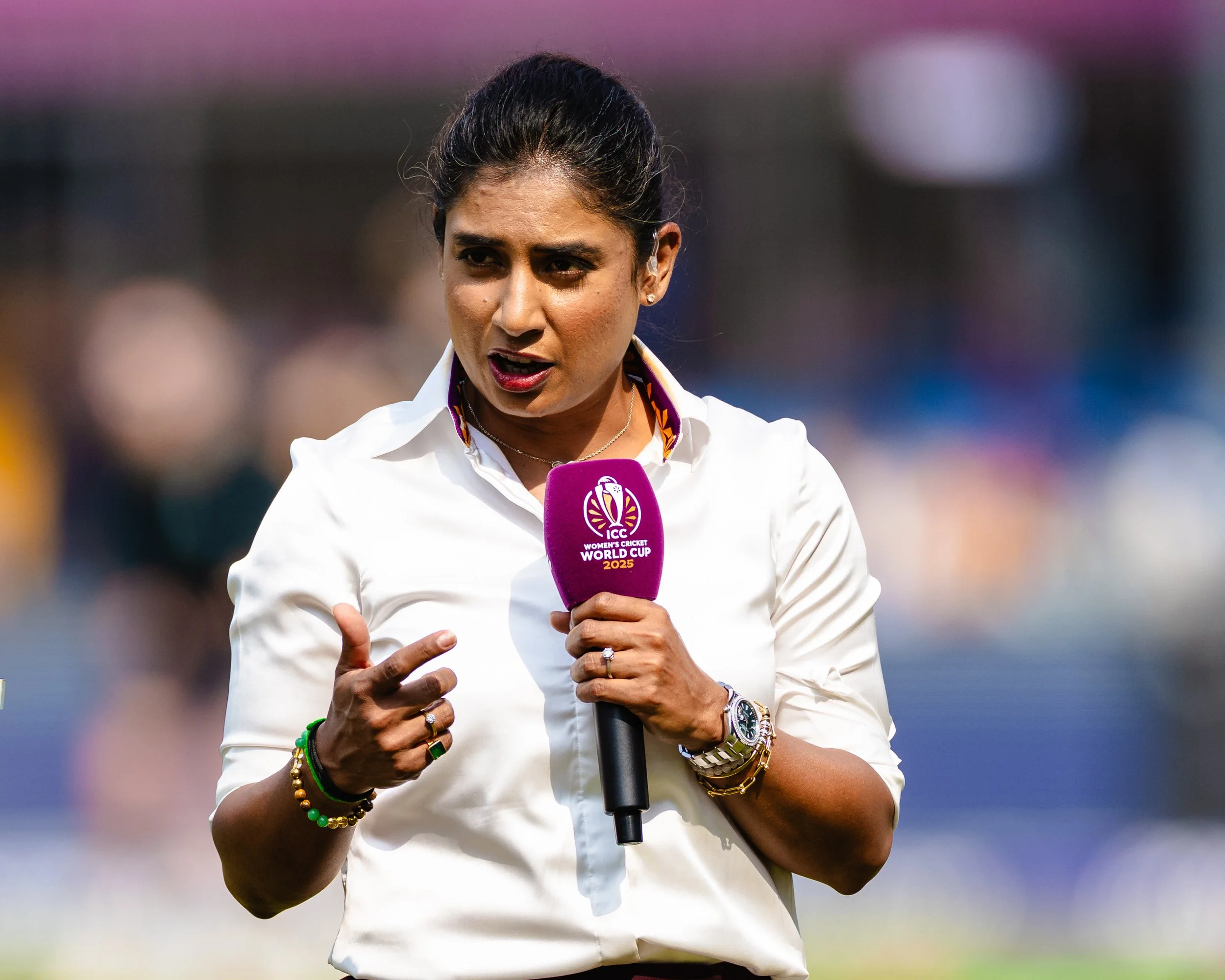 A woman speaking into a microphone during a cricket event, wearing a white shirt and accessorized with jewelry, during daytime on a cricket field.