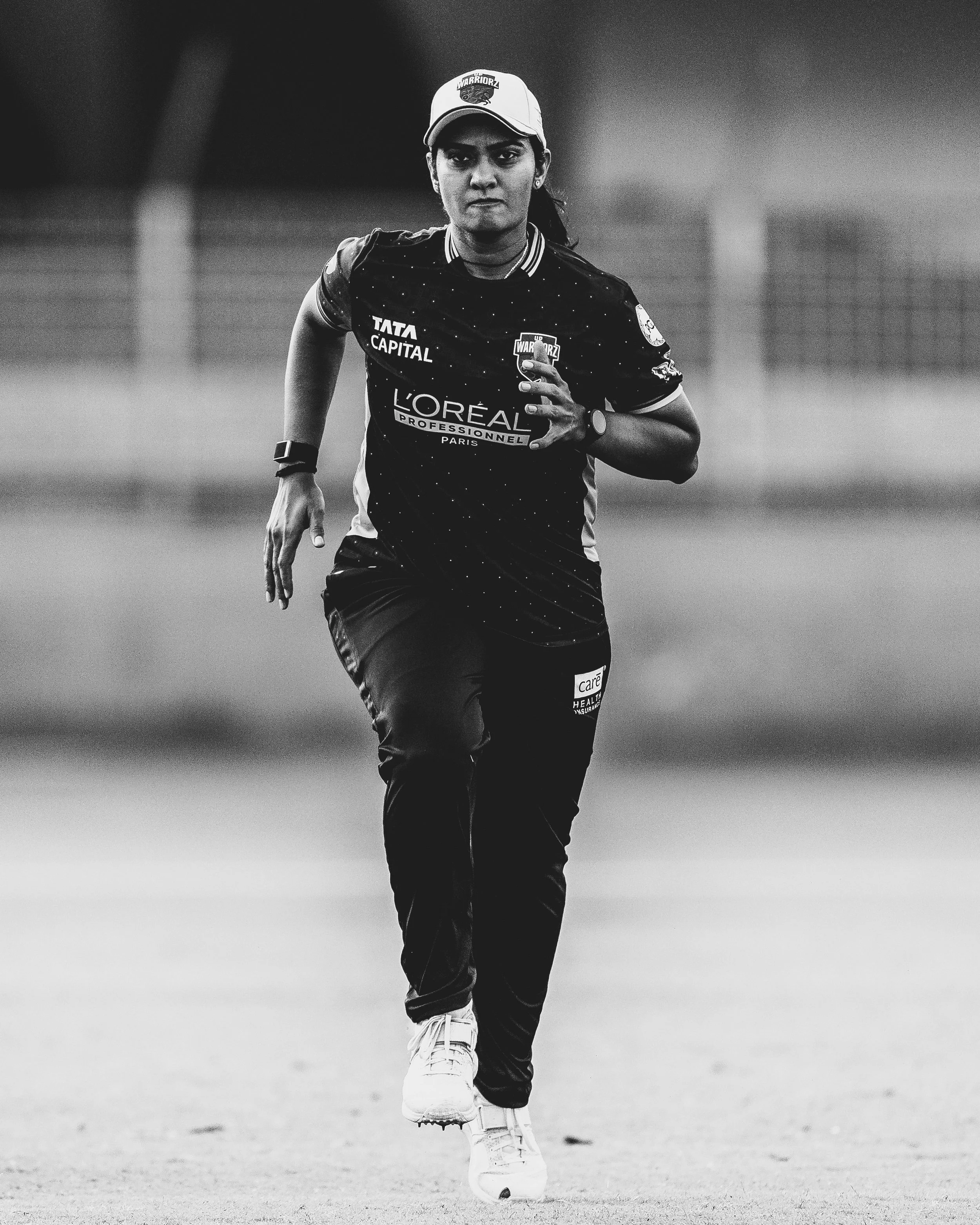 Black and white photograph of a woman running on a field, wearing a sports uniform with sponsorship logos, a cap, and athletic shoes, with a focused expression.