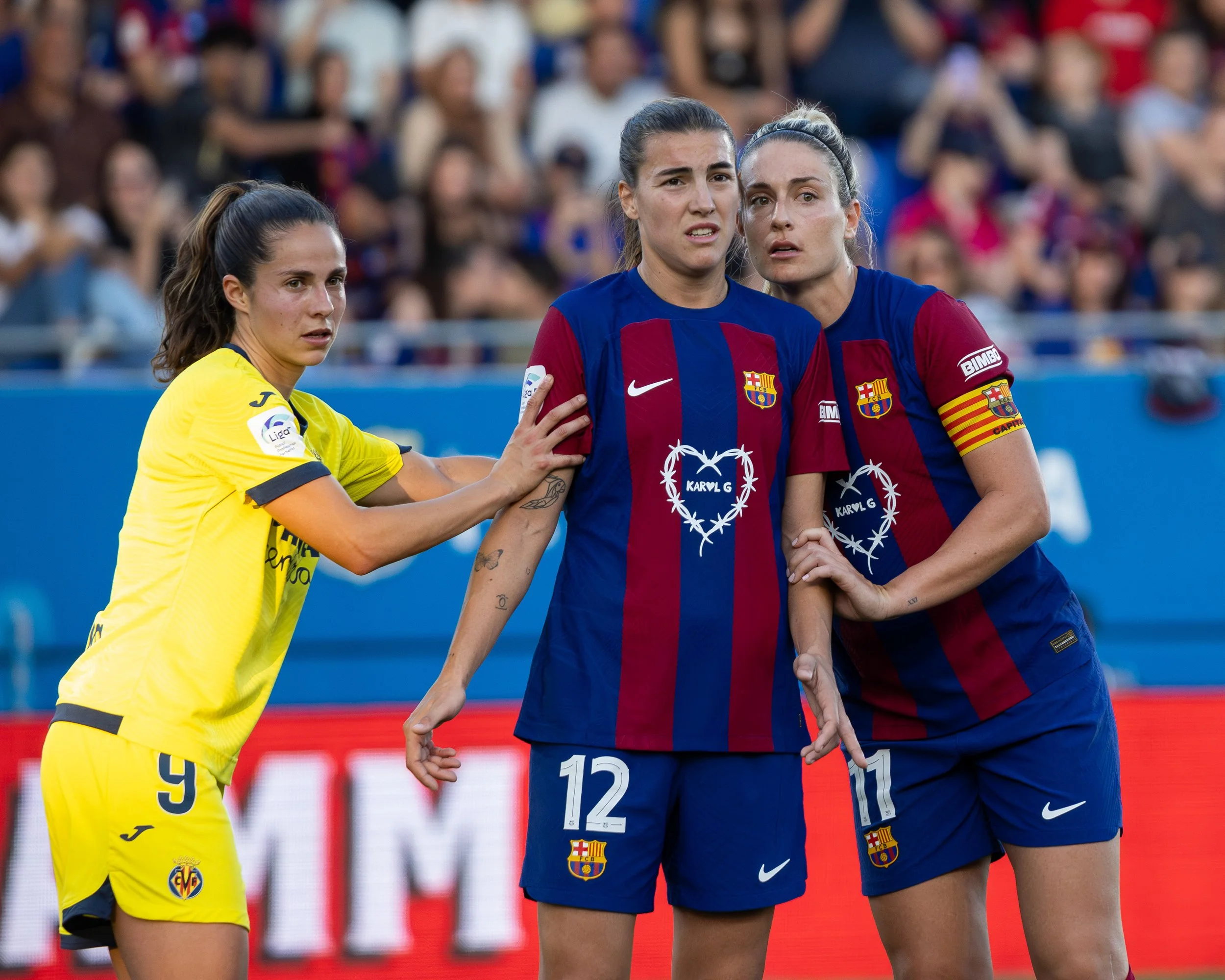 Three women soccer players, two in blue FC Barcelona jerseys and one in yellow team jersey, standing close together on the field during a match with a crowd in the background.