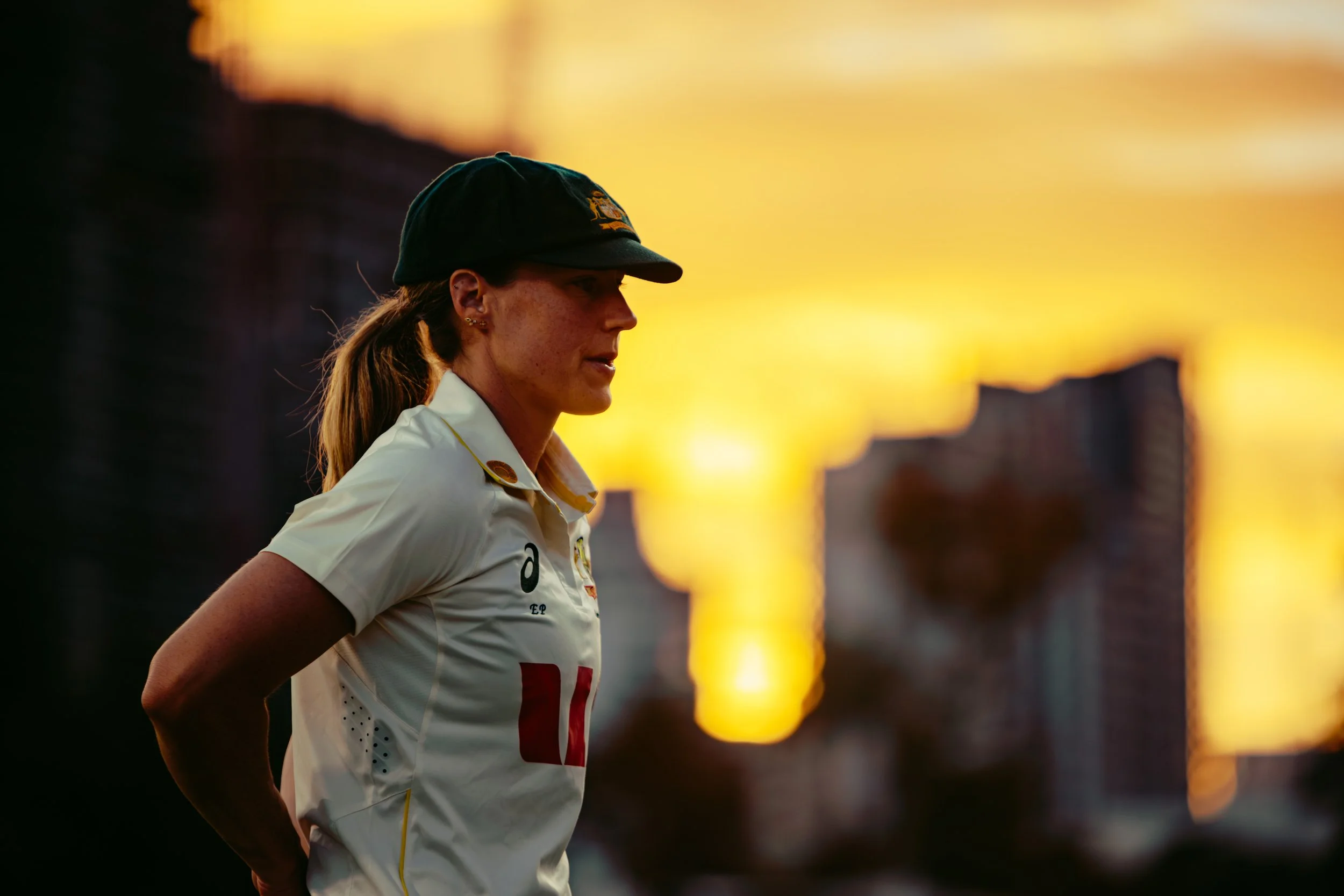 A woman in a sports uniform, wearing a cap, is standing against a vibrant sunset background.