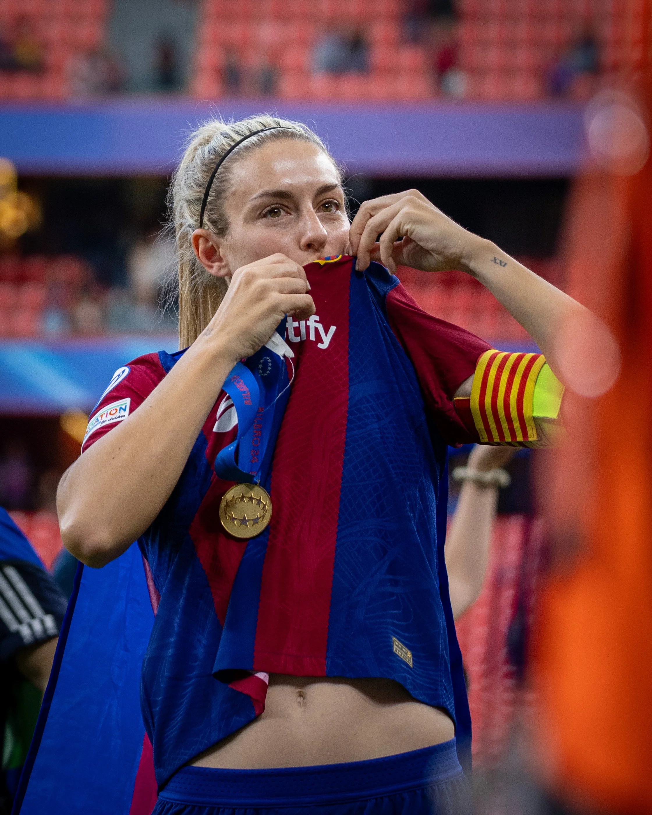 A female athlete with a blonde ponytail and black headband, in a blue and red sports jersey, is pulling up her jersey to reveal a gold medal hanging around her neck.