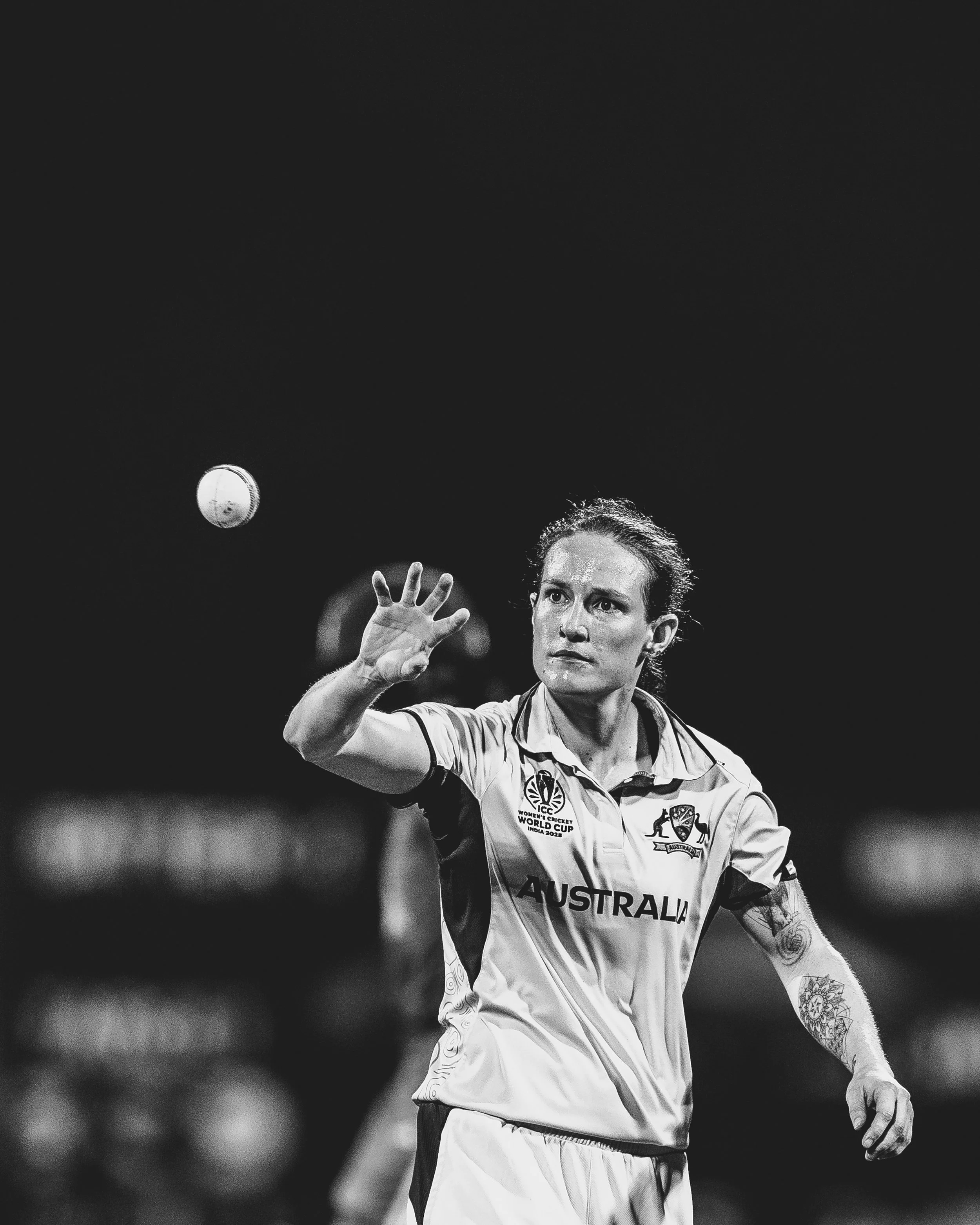 Female cricket player in uniform, with tattoos on left arm, preparing to catch a cricket ball during a match.