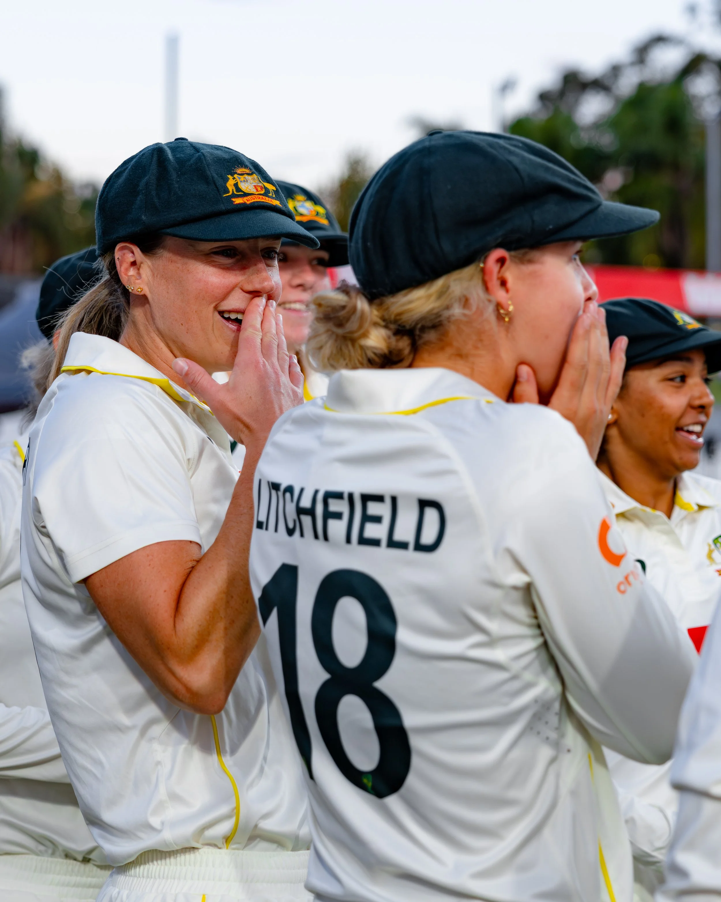 Women wearing black caps and white uniforms, some with visible national emblems, engaged in a team activity or celebration outdoors.