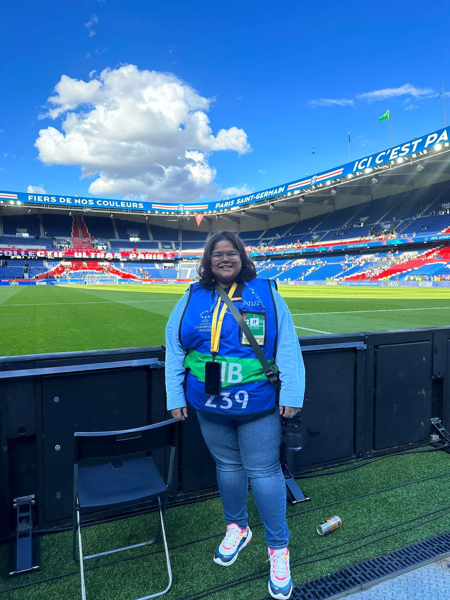A woman in athletic clothing and glasses stands on a soccer field at Paris Saint-Germain stadium, smiling. She wears a medal and an event bib, with a stadium seating area behind her and a blue sky with clouds above.